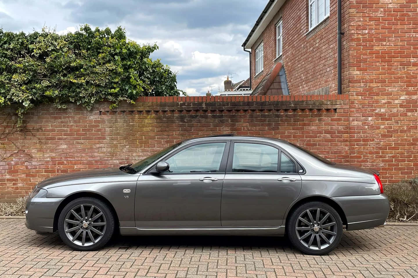 Mg Zt 桌布 壁紙 — 一輛時尚的銀色轎車停放在磚砌車道上，旁邊是紅磚牆和茂密的樹籬，天空多雲。