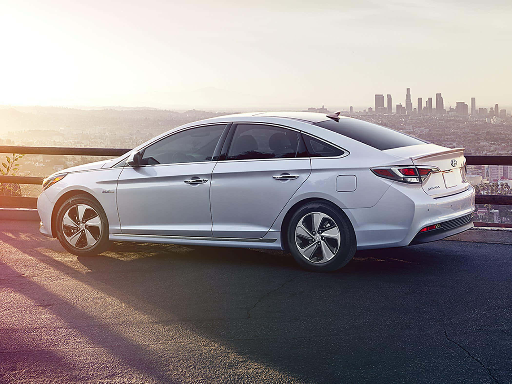 Hyundai Sonata 桌布 壁紙 — 一輛時尚的銀色轎車停泊在山坡上，俯瞰著沐浴在金色日落光芒中的廣闊城市天際線。