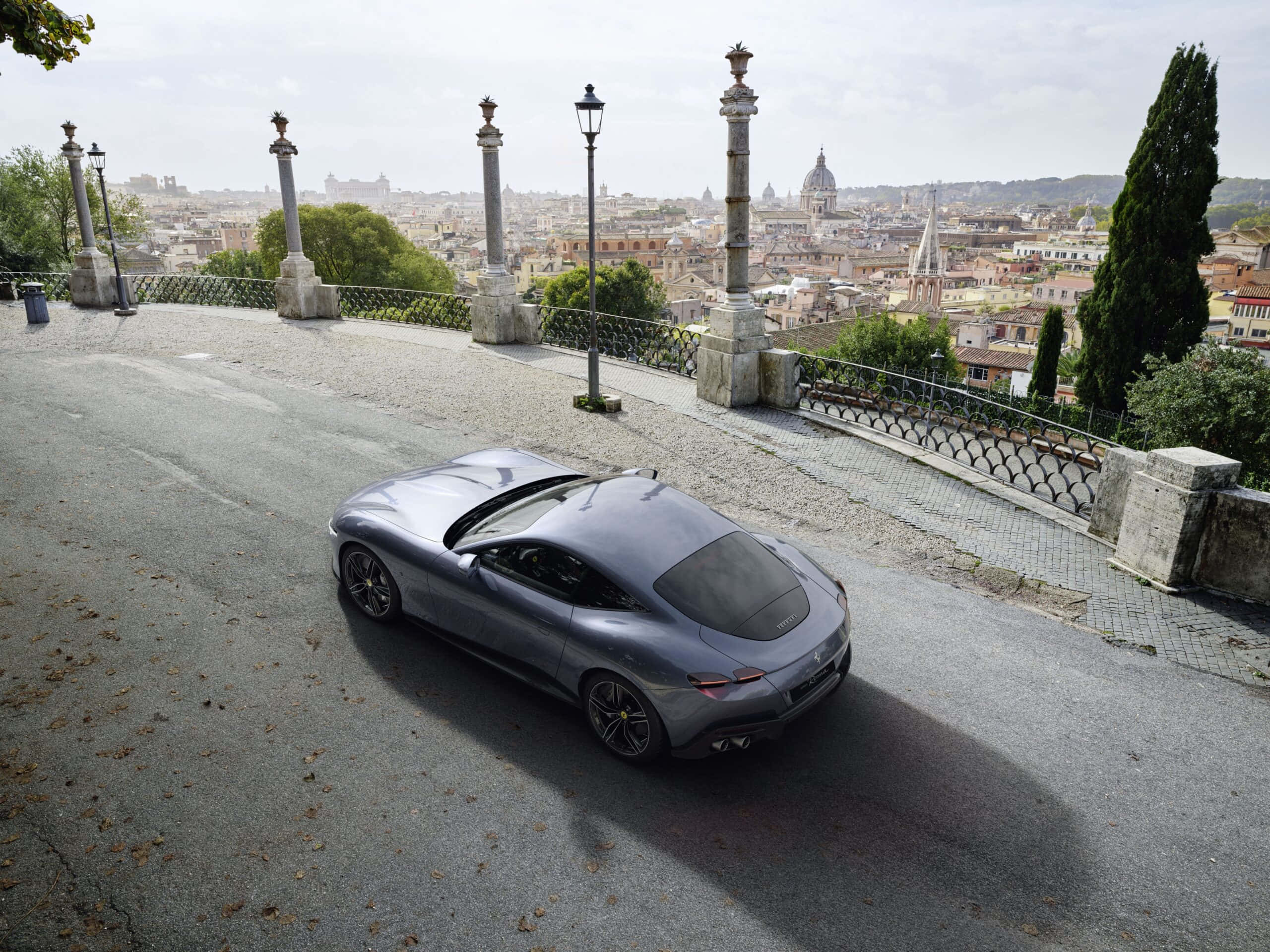 Ferrari Roma 桌布 壁紙 — 一輛時尚的銀色跑車沿著鵝卵石山坡行駛，俯瞰著陰天下一片廣闊的歷史城市景觀。