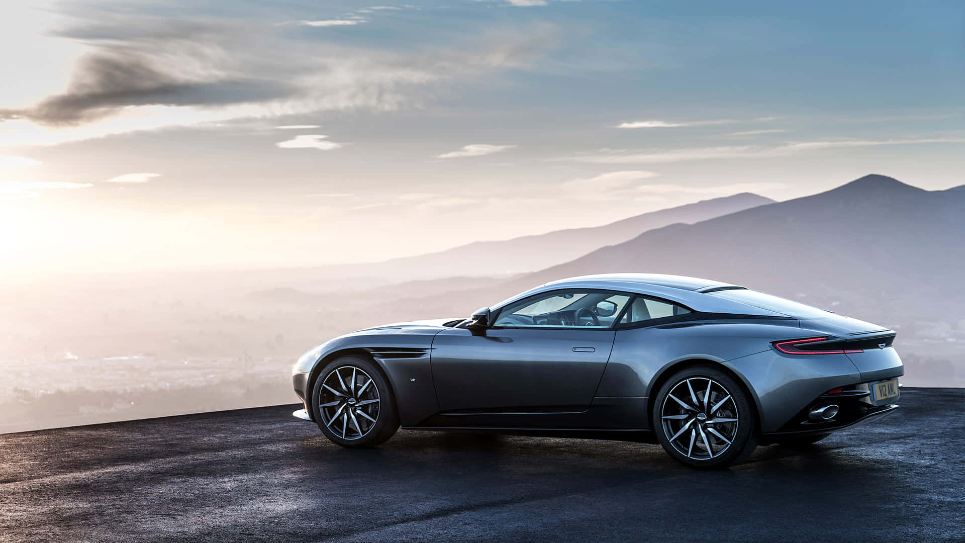 Aston Martin Db11 桌布 壁紙 — 一輛時尚的銀色跑車停在懸崖上，俯瞰日出時遠處的山脈，伴隨著柔和的金色光芒。
