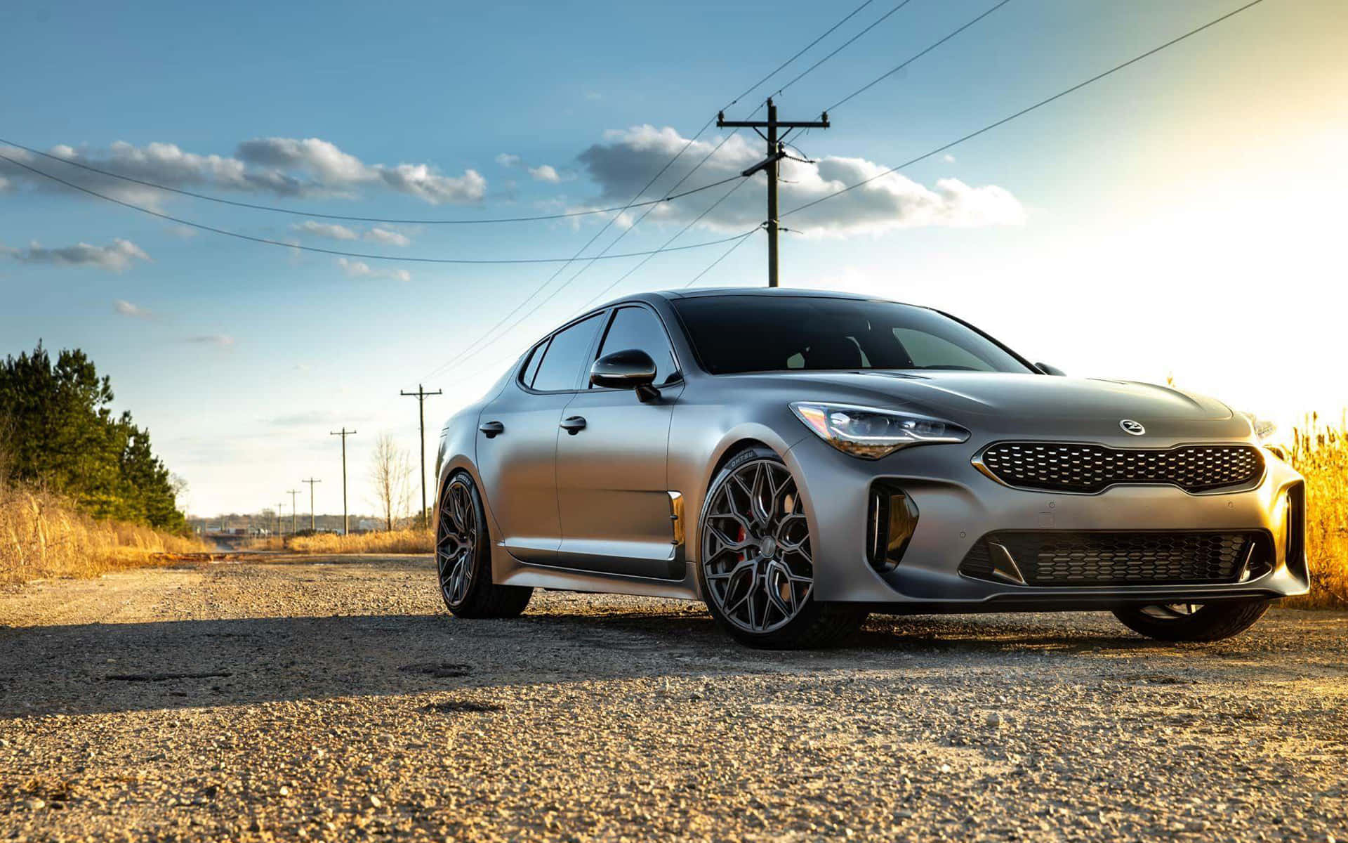 Kia Stinger 桌布 壁紙 — 一輛時尚的銀色跑車停在碎石路上，上方是晴朗藍天，點綴著幾朵白雲。