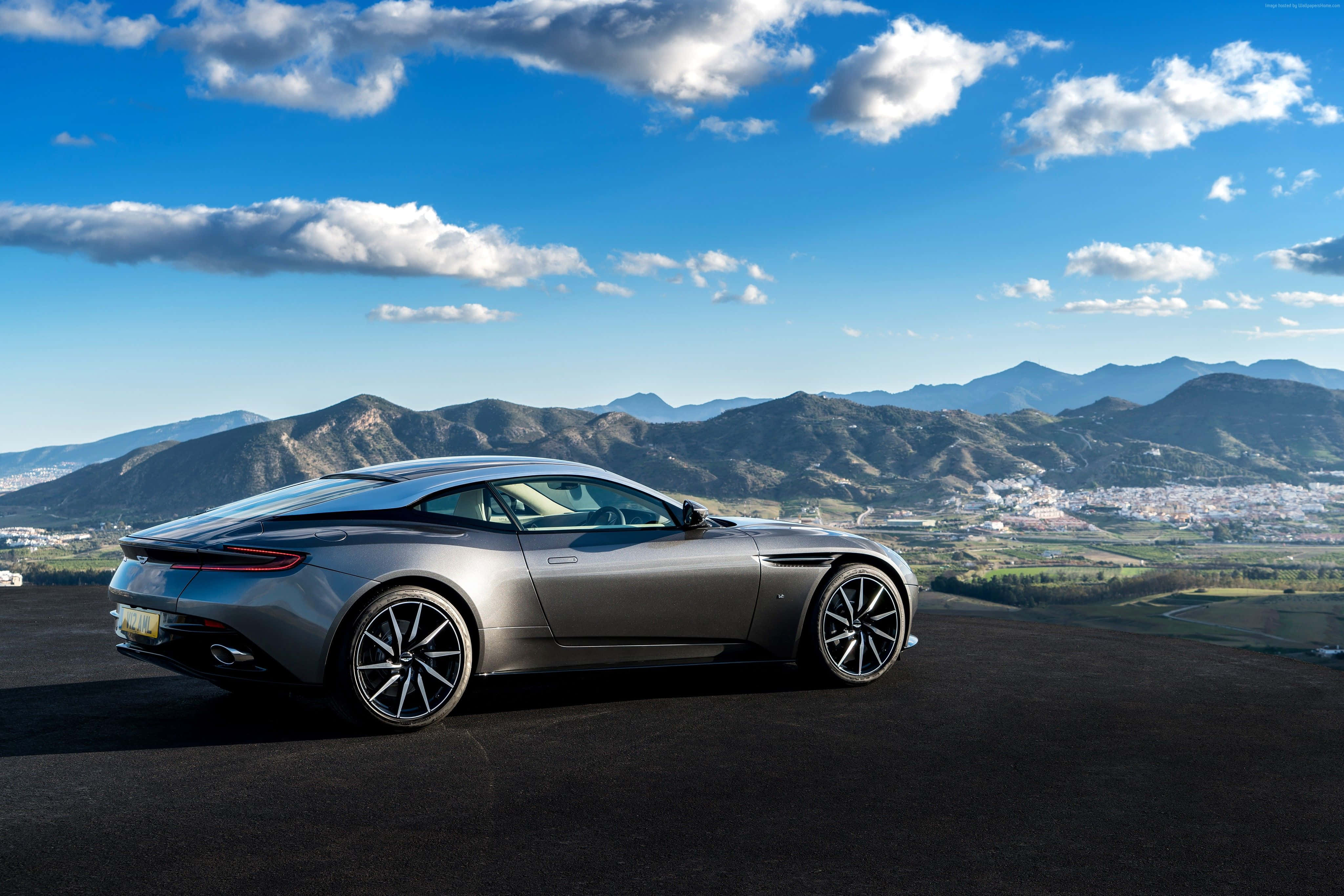 Aston Martin Db11 桌布 壁紙 — 一架時尚的銀色跑車停在山頂上，俯瞰山谷小鎮，天空湛藍，白雲朵朵。