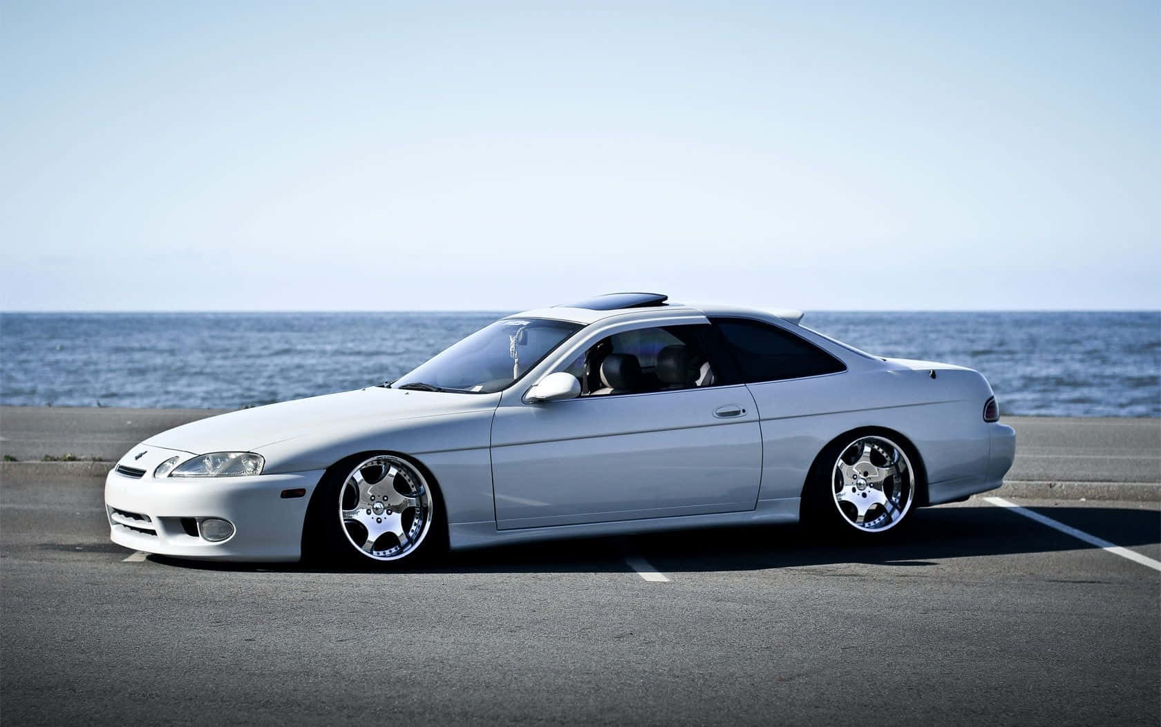 Lexus Sc 300 桌布 壁紙 — 一輛時尚的銀色運動轎跑車配備訂製輪圈，停在沿海公路上，旁邊是海洋，天空清澈湛藍。
