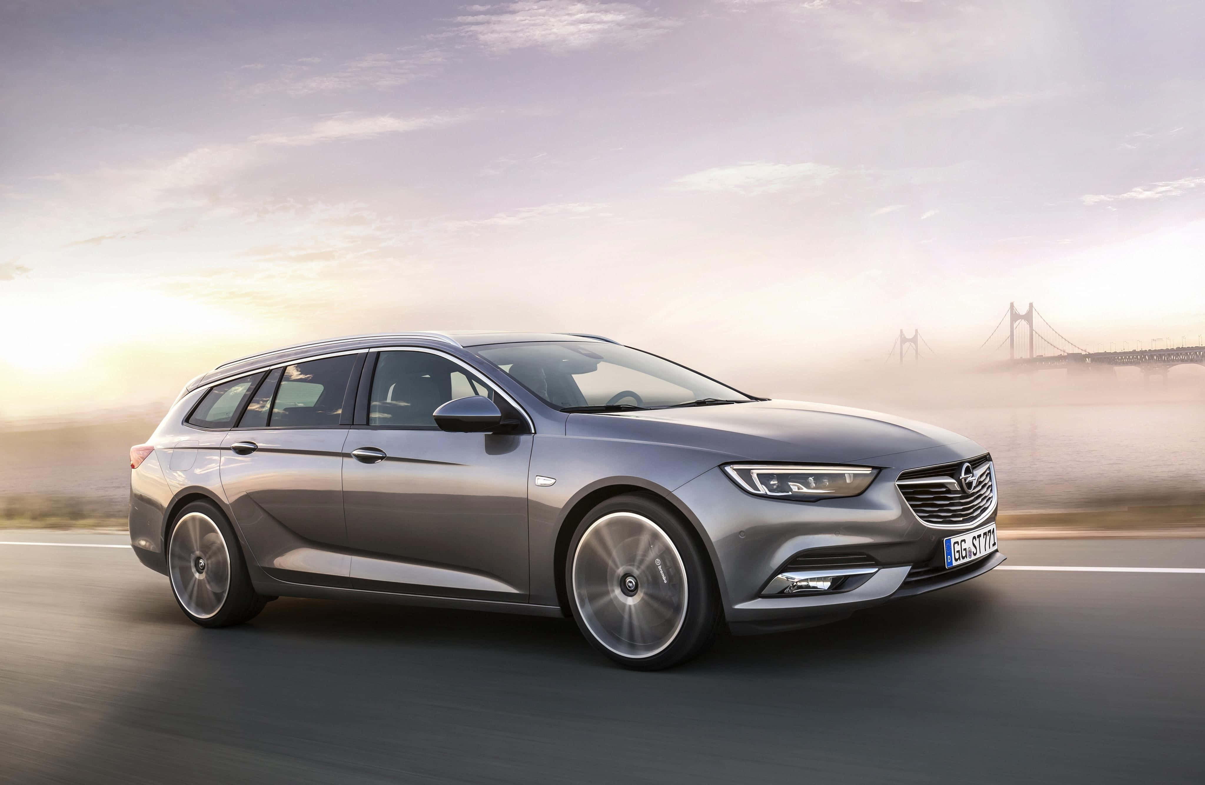 Opel Insignia 桌布 壁紙 — 一輛時尚的銀色旅行車沿著海岸公路在日出時行駛，遠處有一座霧濛濛的橋樑。