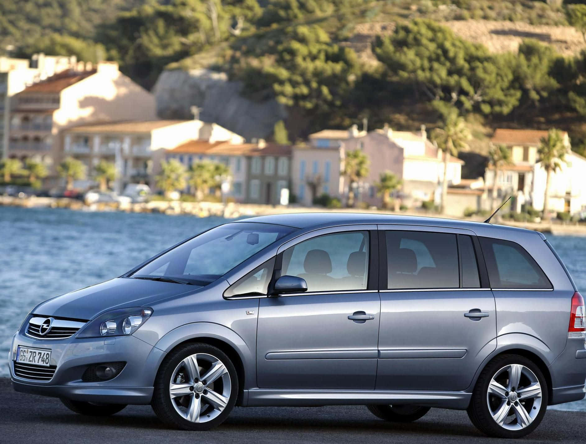 Opel Zafira 桌布 壁紙 — 一輛時尚的銀色旅行車停在海濱，背景是地中海風格的房屋和綠色山丘。