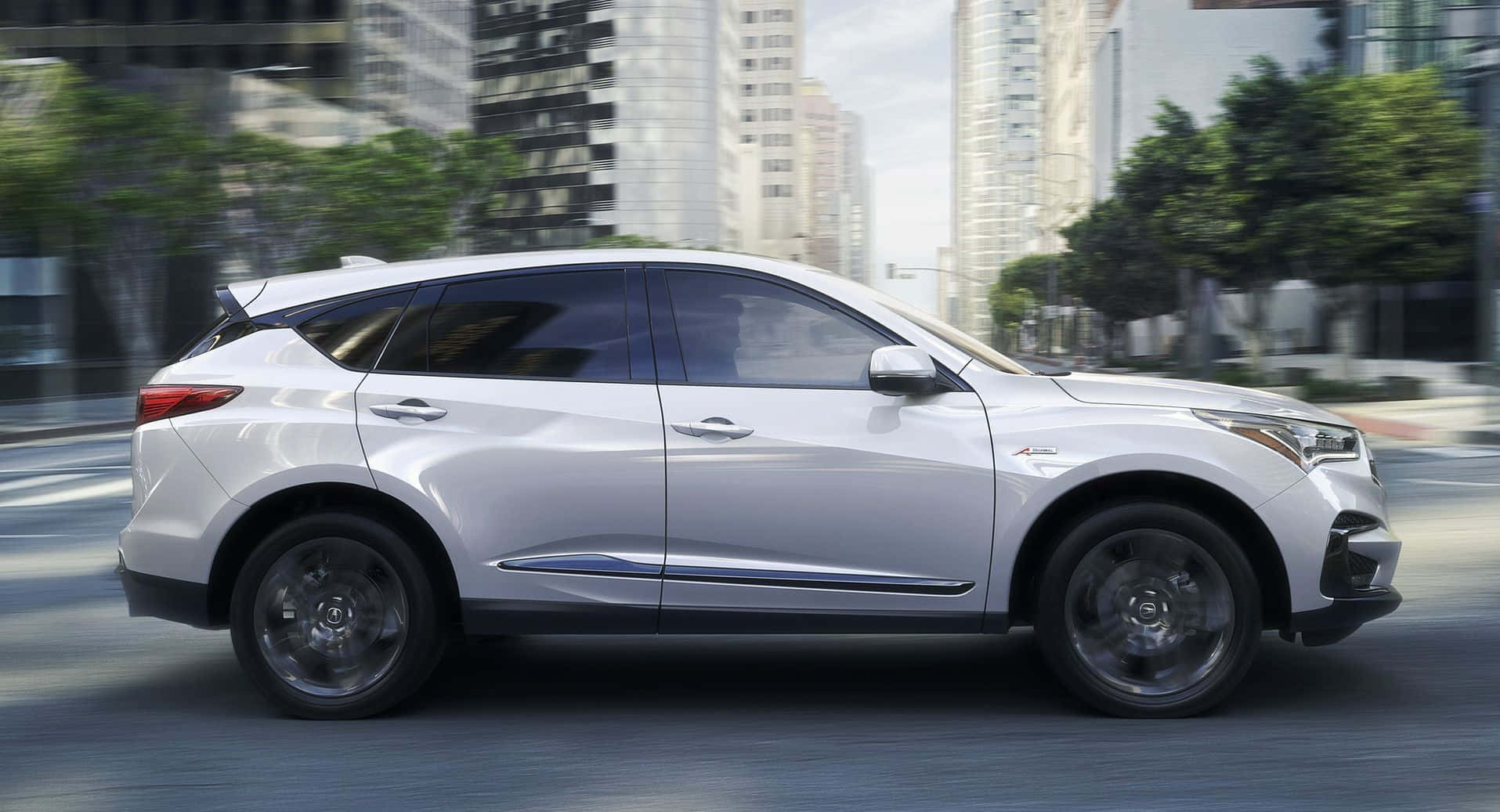 Acura Rdx 桌布 壁紙 — 一輛時尚的銀色SUV在現代城市街道上飛馳，背景是模糊的摩天大樓和綠樹。