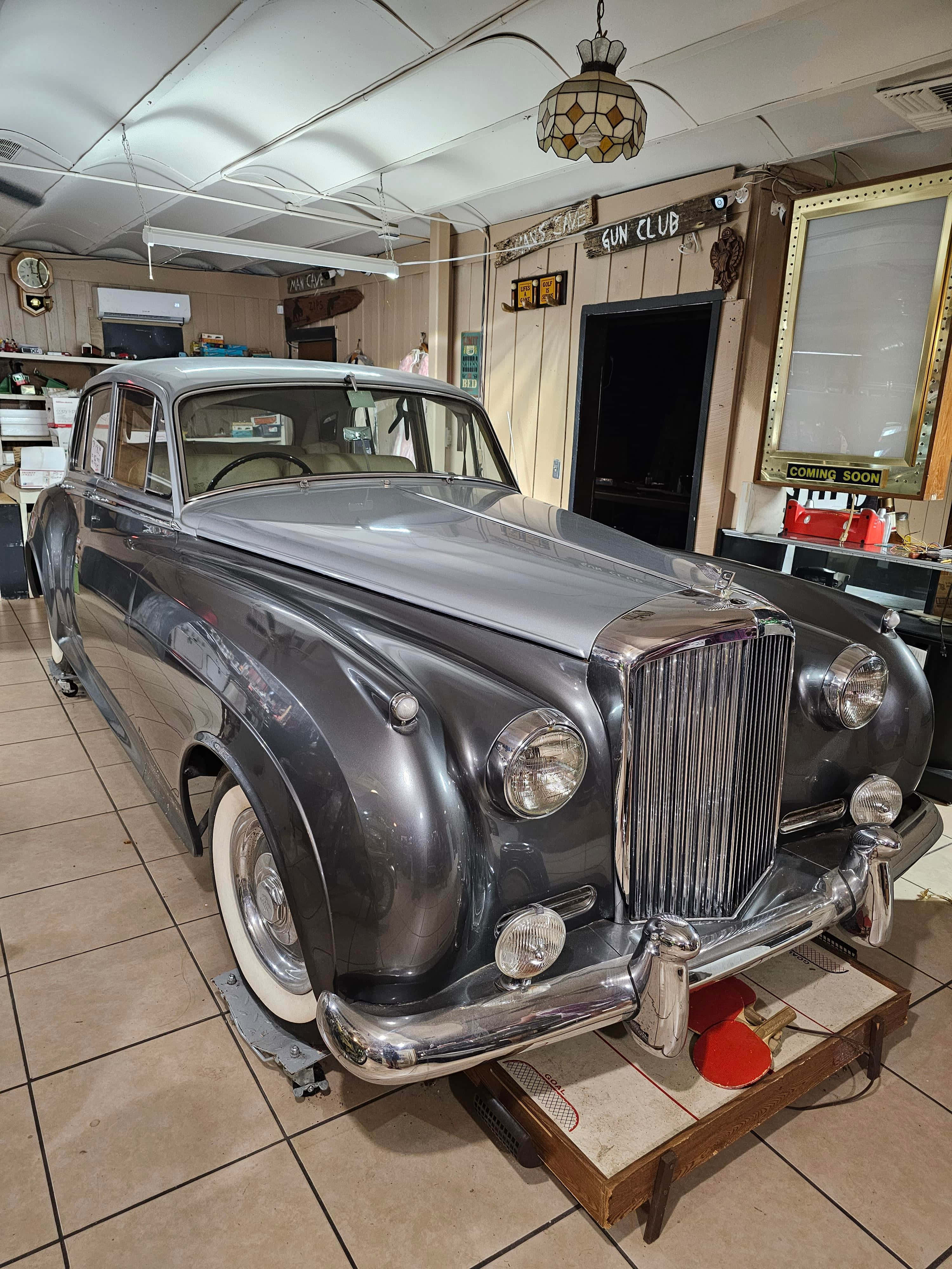 Bentley S2 桌布 壁紙 — 一輛時尚的銀色復古車在鋪有瓷磚的車庫中，在溫暖的燈光下閃閃發光，車庫裝飾著復古標誌和裝飾品。