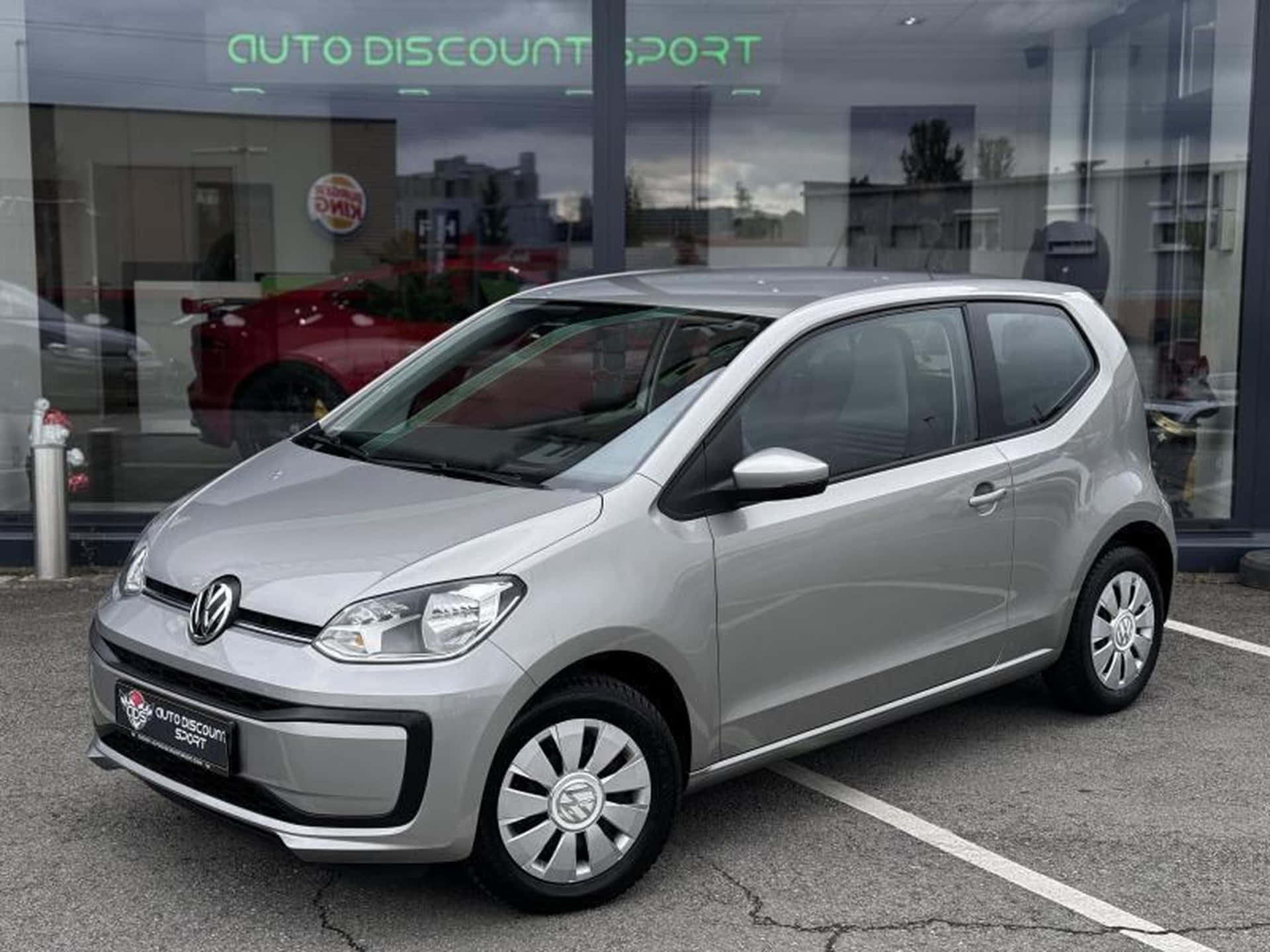  壁紙 — 一輛時尚的銀色Volkswagen up!停放在玻璃窗反射多雲天空和內部紅色汽車的汽車經銷商前。