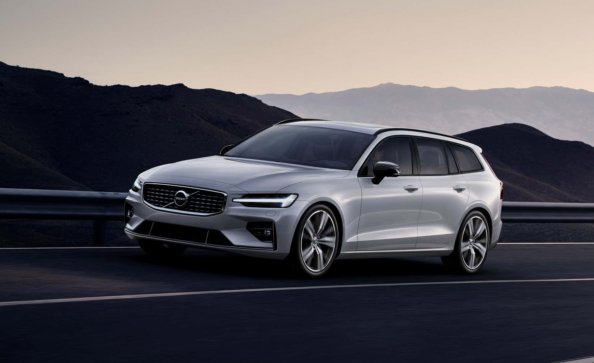 Volvo V60 桌布 壁紙 — 一台時尚的銀色富豪旅行車在黃昏時分沿著山區高速公路行駛，環境燈光柔和。