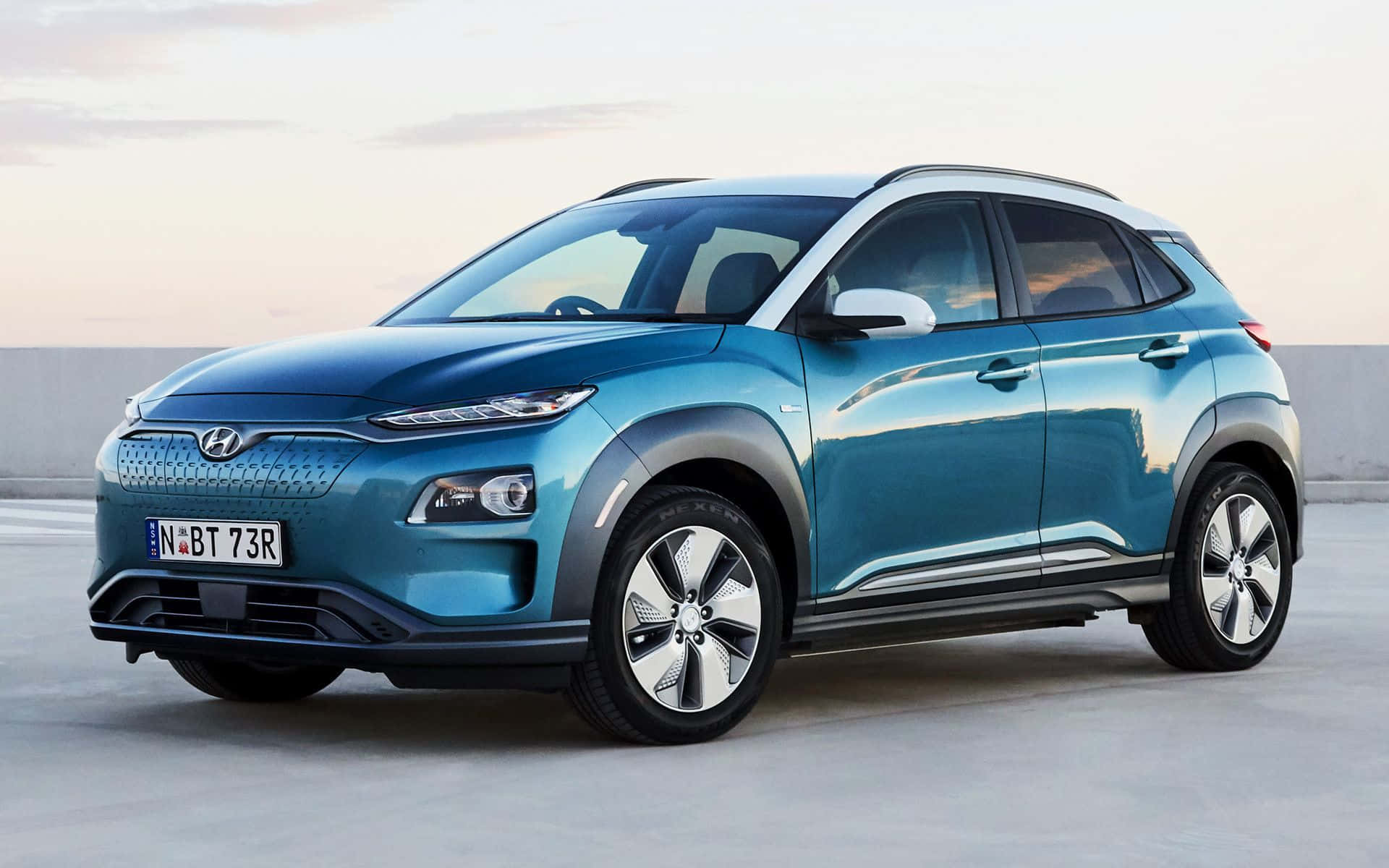 Hyundai Kona 桌布 壁紙 — 一輛時尚的青綠色Hyundai Kona電動SUV停放在混凝土表面上，在柔和的暮色天空下。