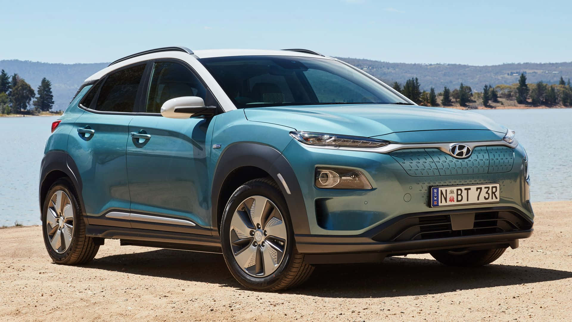 Hyundai Kona 桌布 壁紙 — 一輛時尚的藍綠色現代Kona電動SUV停泊在沙灘上，旁邊是平靜的水面，背後是樹木茂密的山丘，上方是明亮的藍天。