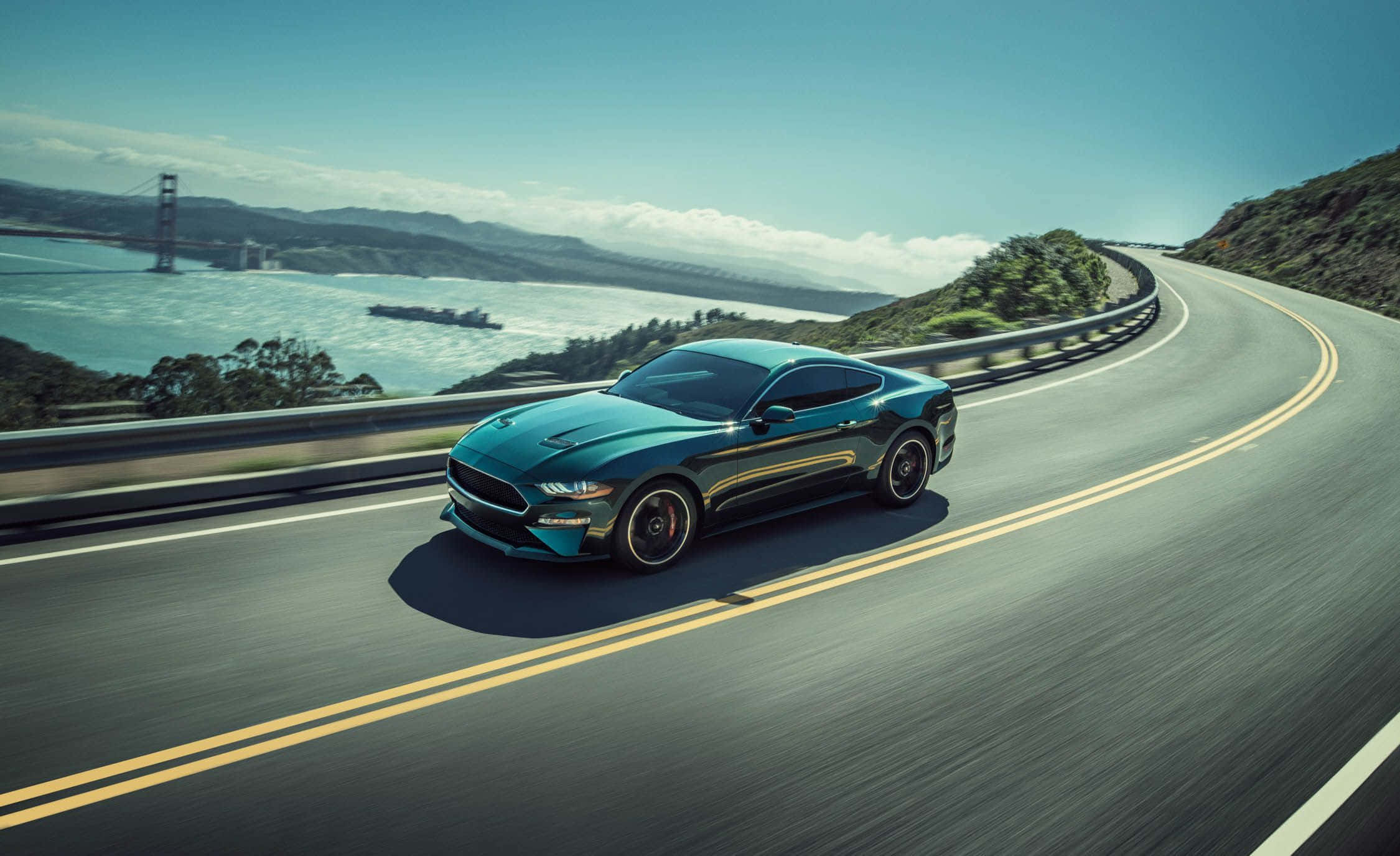 Ford Mustang Bullitt 桌布 壁紙 — 一架時尚的青綠色Mustang沿著海岸高速公路奔馳，遠方的金門大橋在晴朗的藍天下清晰可見。
