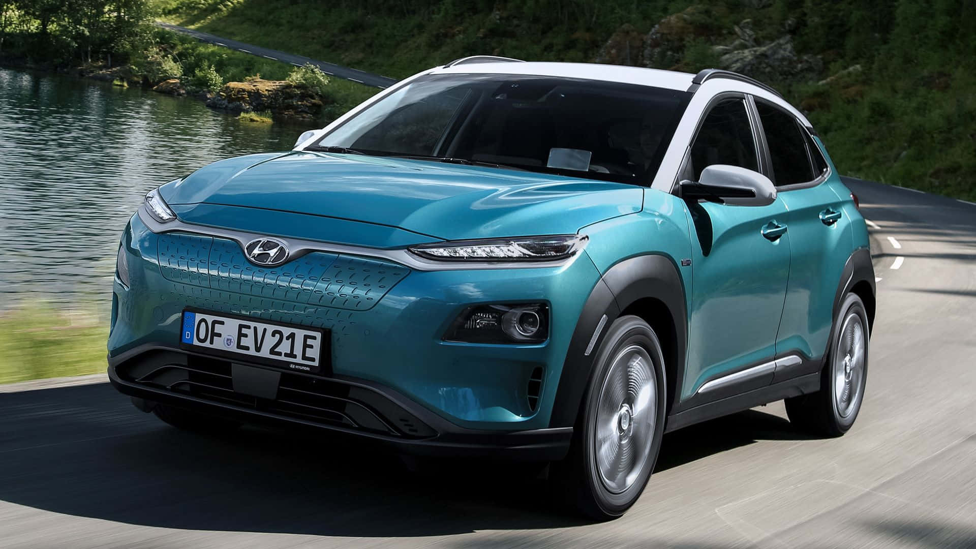 Hyundai Kona 桌布 壁紙 — 一輛時尚的藍綠色Hyundai Kona EV在明亮的日光下沿著蜿蜒的道路行駛，旁邊是一條平靜的河流。