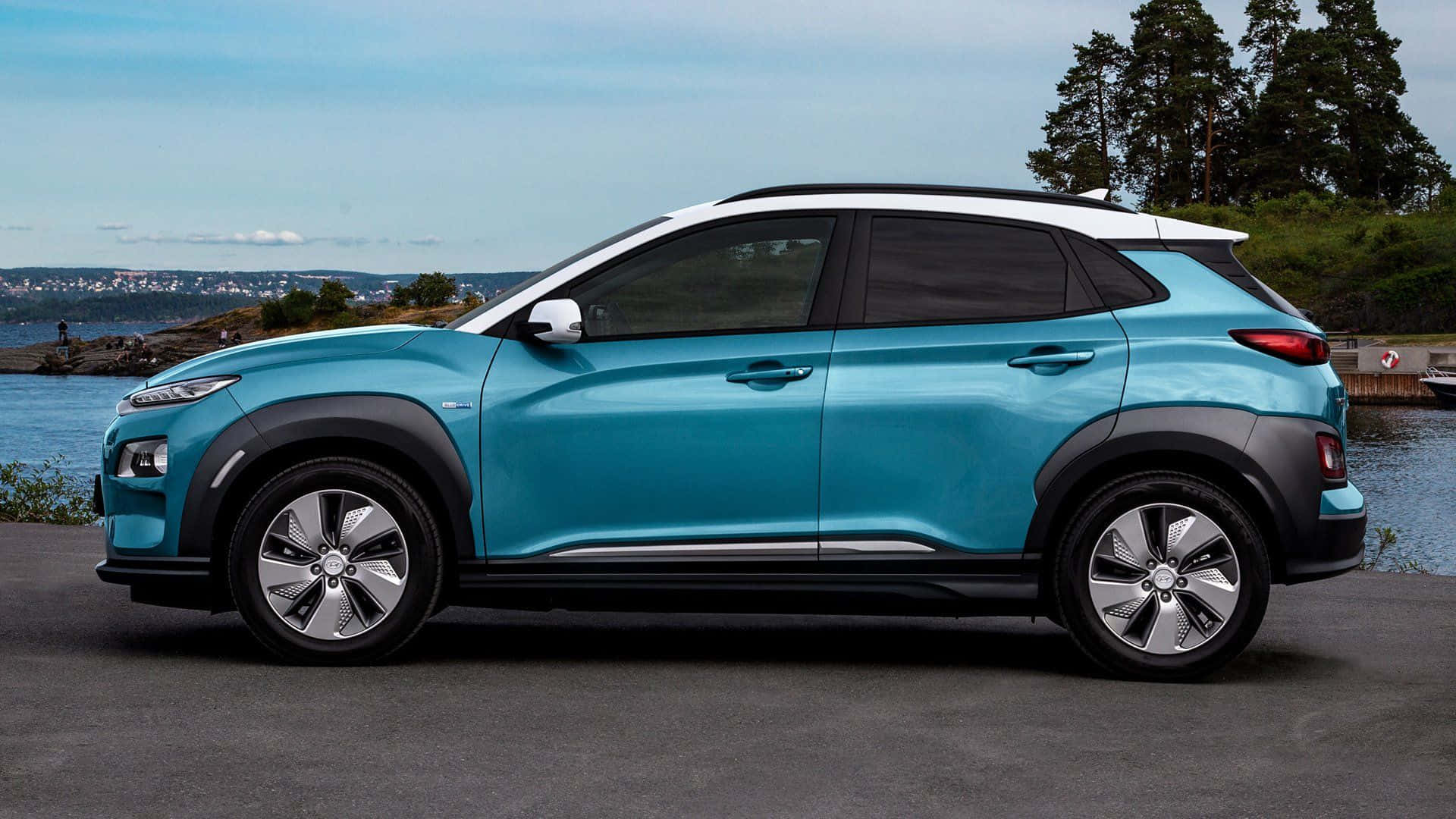 Hyundai Kona 桌布 壁紙 — 一輛時尚的藍綠色SUV停泊在海濱，旁邊有樹木和遠處的城市天際線，天空晴朗。