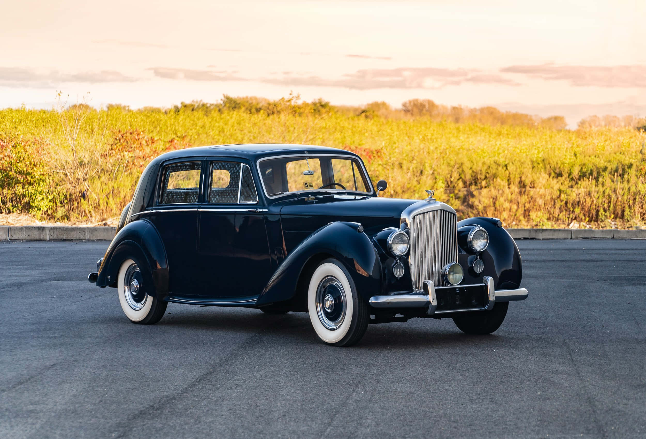 Bentley Mark Vi 桌布 壁紙 — 一架時尚的復古藍色 Bentley 轎車配備白色側壁輪胎，在金色夕陽下於瀝青場地上閃閃發光。