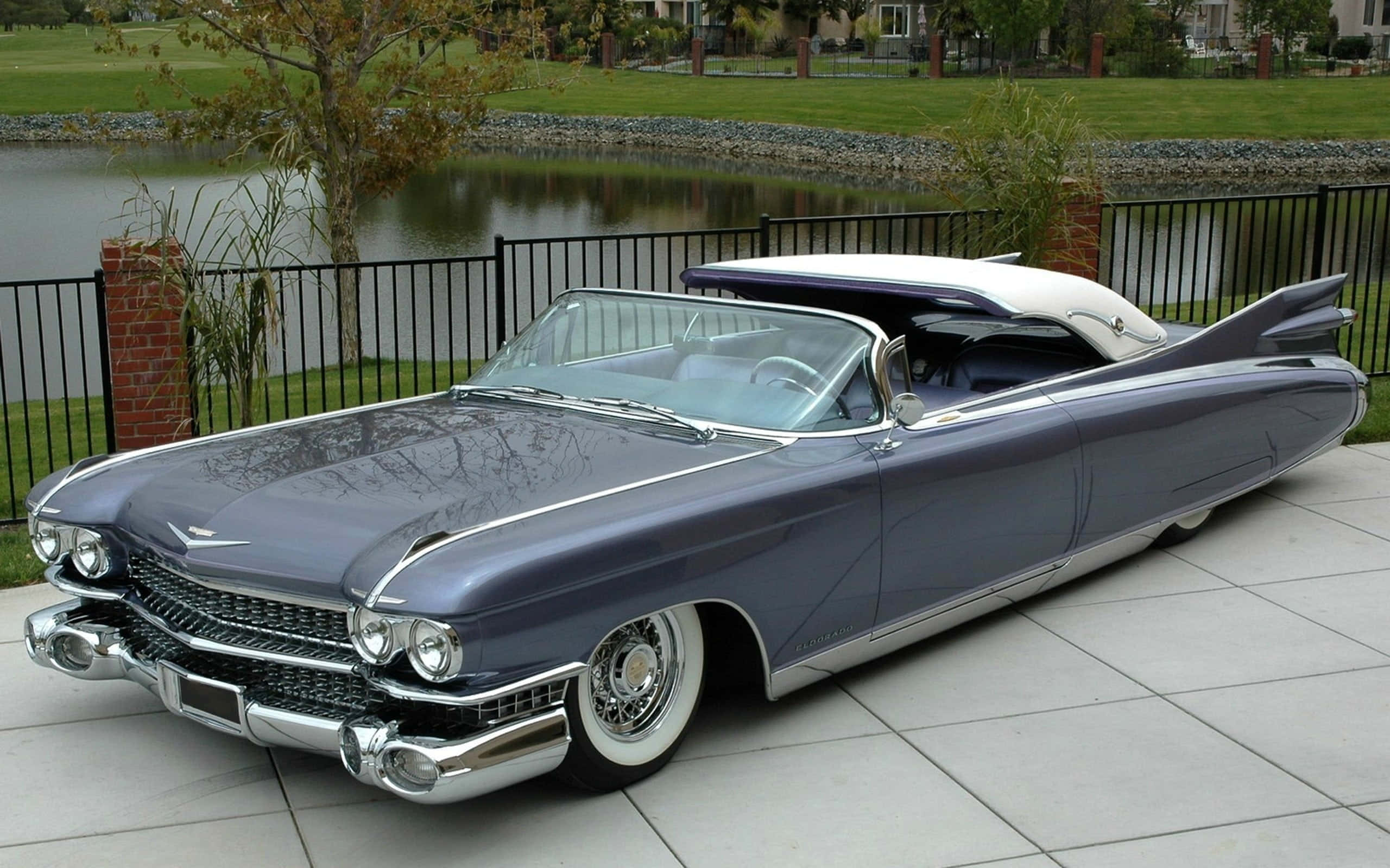Cadillac Eldorado 桌布 壁紙 — 一輛時尚的復古藍色敞篷車，帶有鍍鉻裝飾，停在瓷磚露台上，旁邊是黑色圍欄和寧靜的池塘。