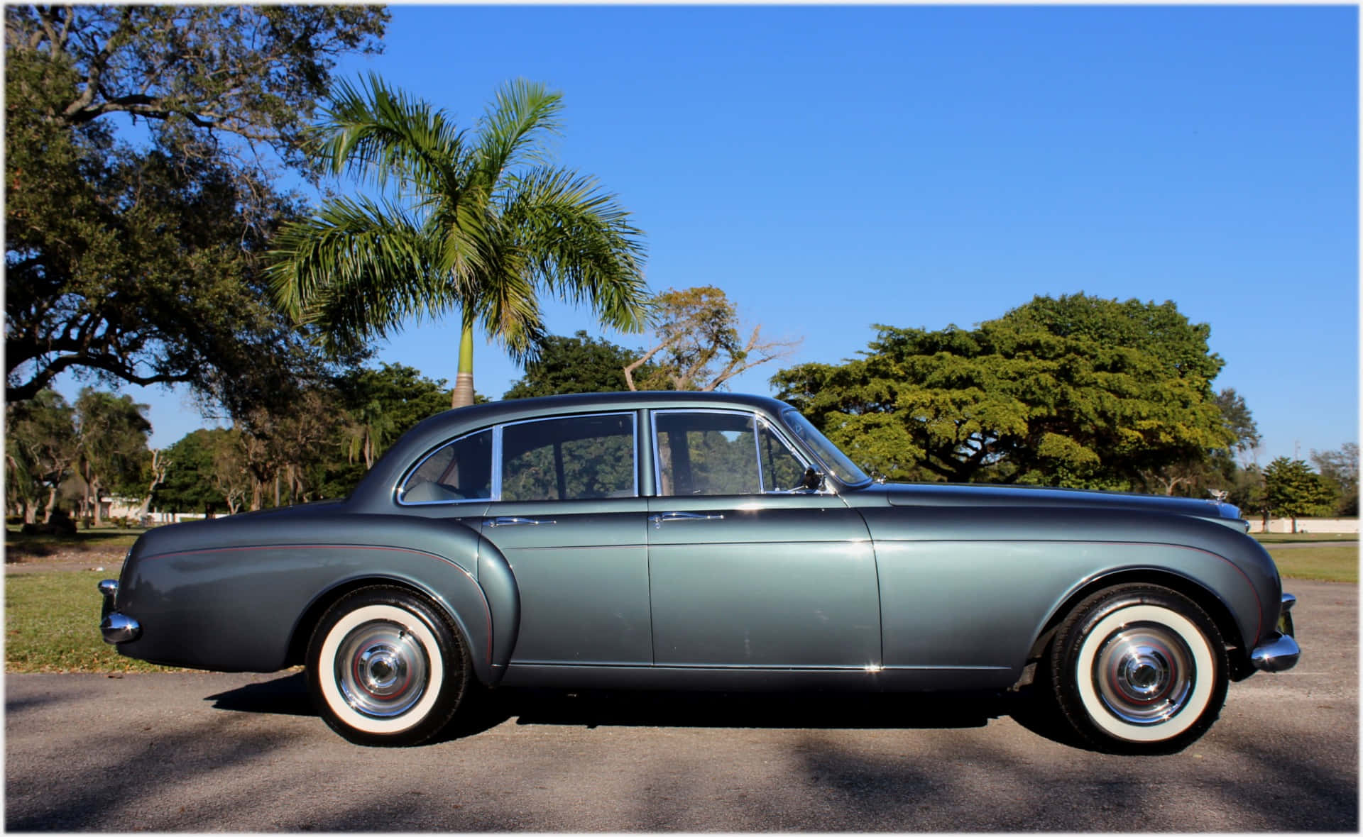 Bentley S2 桌布 壁紙 — 一輛時尚的復古青綠色轎車配備白色側壁輪胎，在明媚陽光下與茂密的棕櫚樹和綠色植物旁閃閃發光。