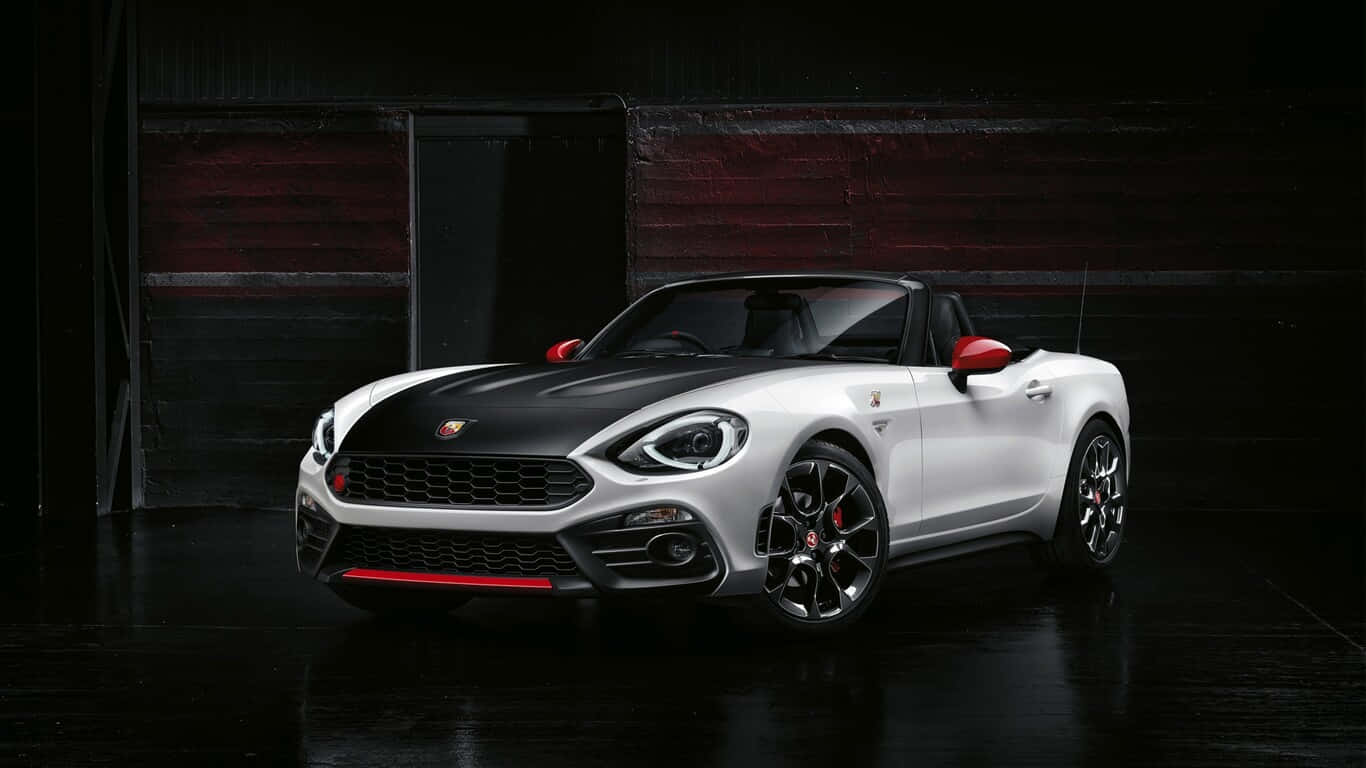 Fiat 124 Spider 桌布 壁紙 — 一輛時尚的白色和黑色敞篷跑車，帶有紅色點綴，在黑暗的工業車庫中於攝影棚燈光下閃閃發光。