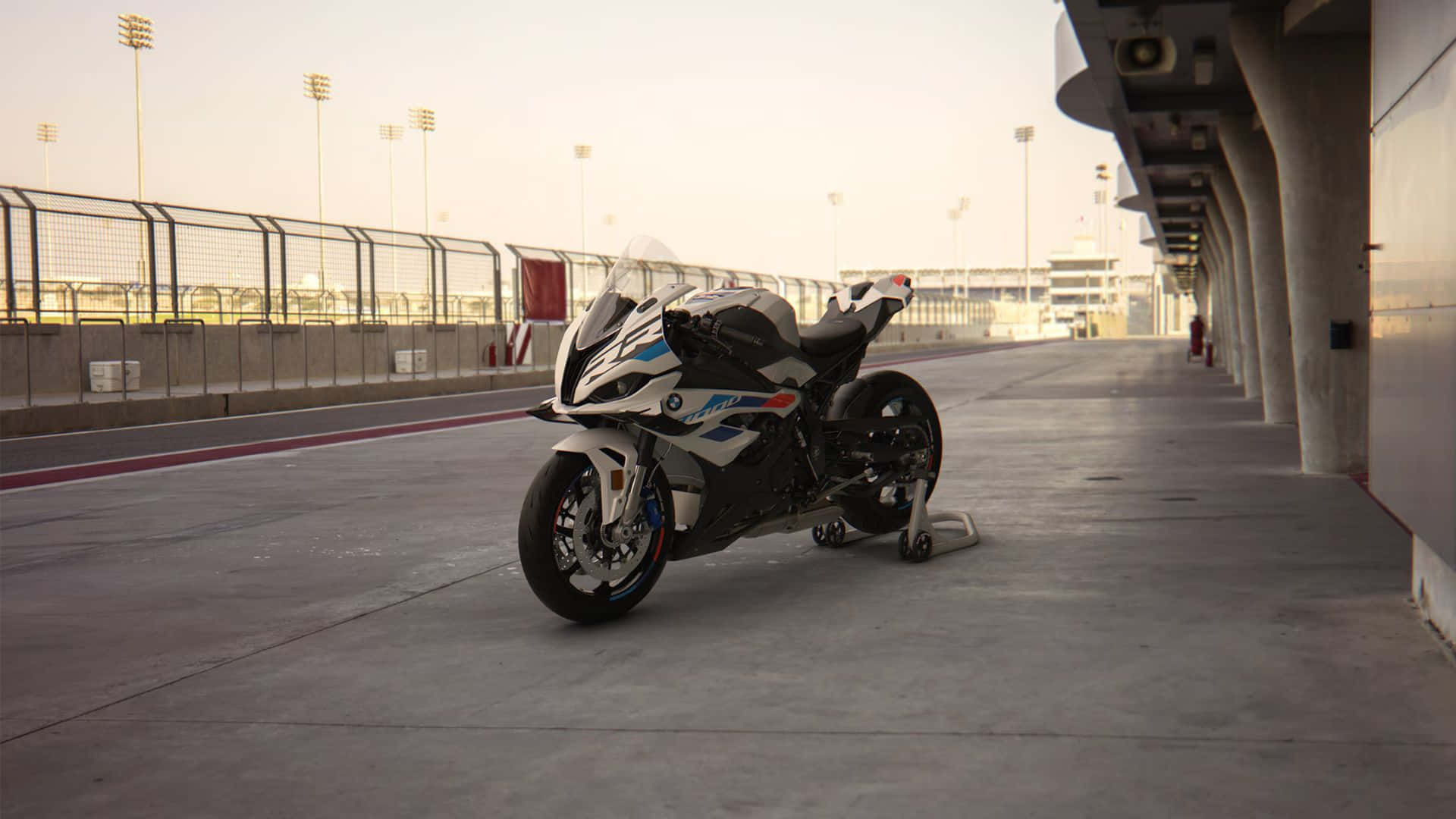  壁紙 — 一架時尚的白色和藍色BMW運動機車，在溫暖的黃金時段光線下，停泊在賽車場的混凝土維修區車道上。