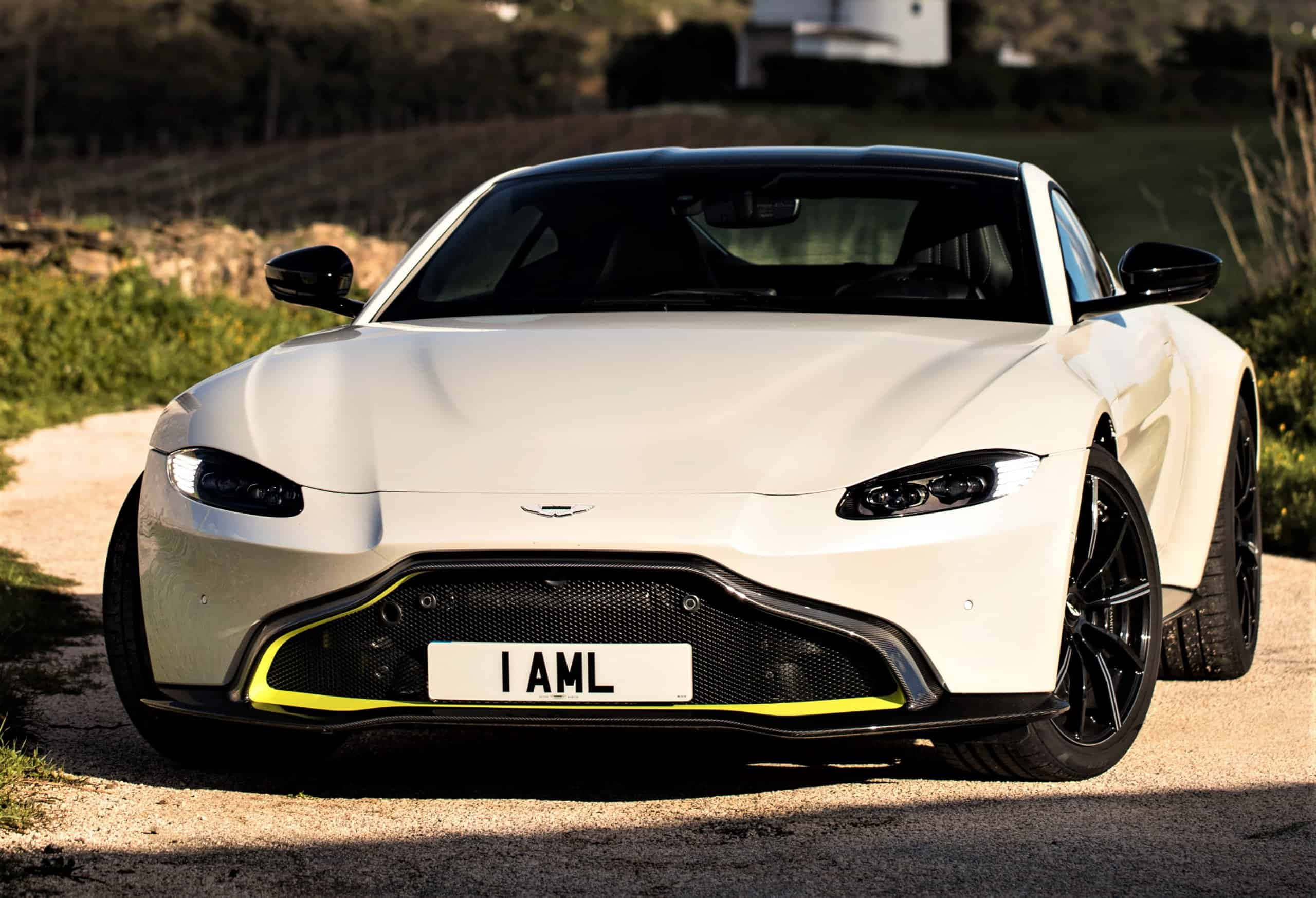 Aston Martin Vantage 桌布 壁紙 — 一輛時尚的白色Aston Martin跑車，帶有黃色點綴，停泊在碎石路上，周圍綠樹成蔭。