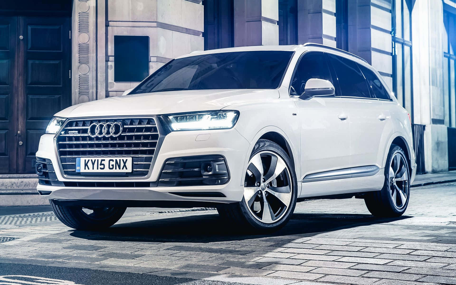Audi Q7 桌布 壁紙 — 一輛時尚的白色奧迪Q7 SUV停泊在鵝卵石街道上，前方是帶有藍色玻璃的現代建築。