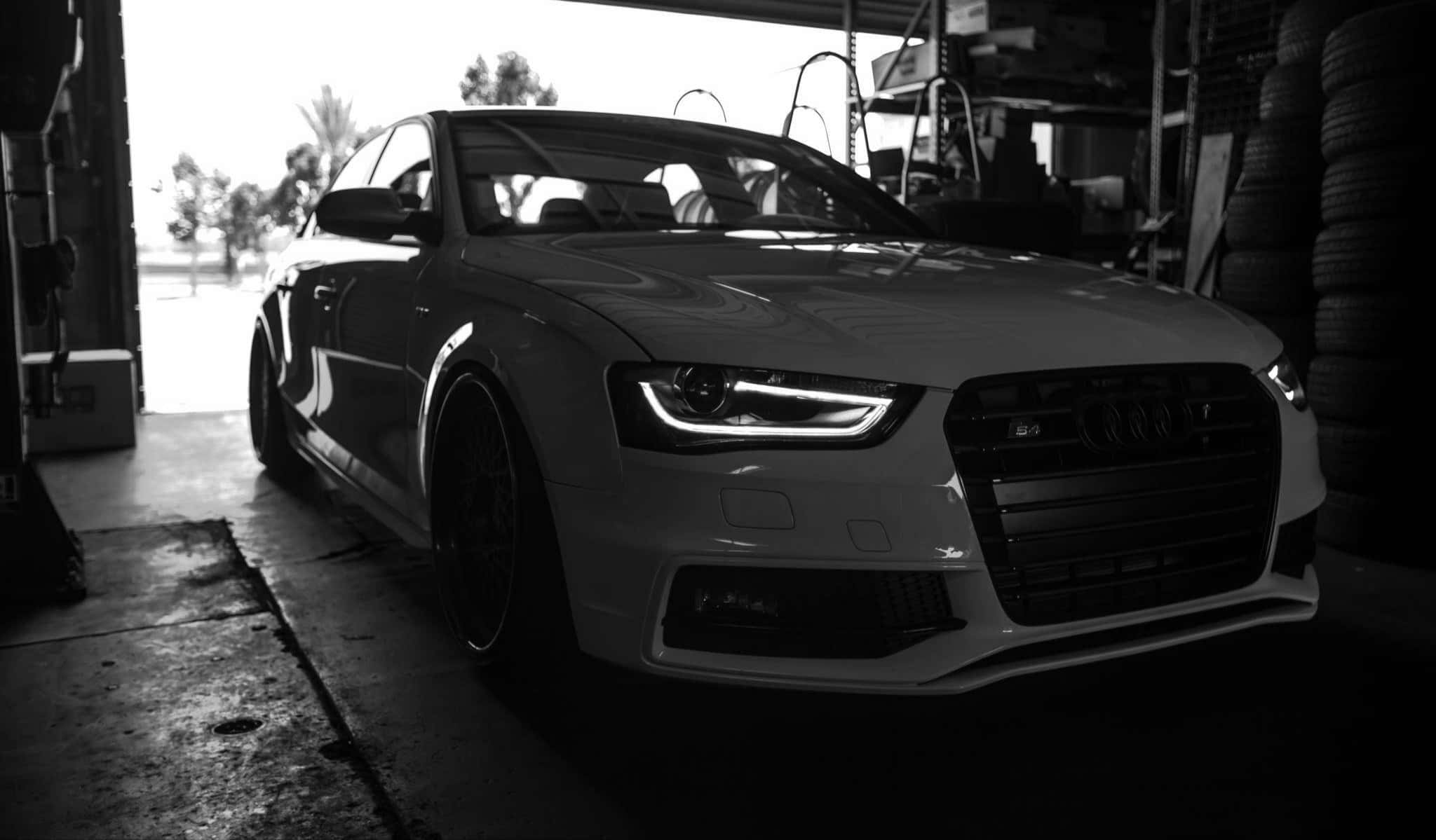 Audi A4 桌布 壁紙 — 一輛時尚的白色奧迪跑車，車頭燈閃閃發光，戲劇性地停在光線昏暗的車庫中，周圍堆滿了輪胎和工具。
