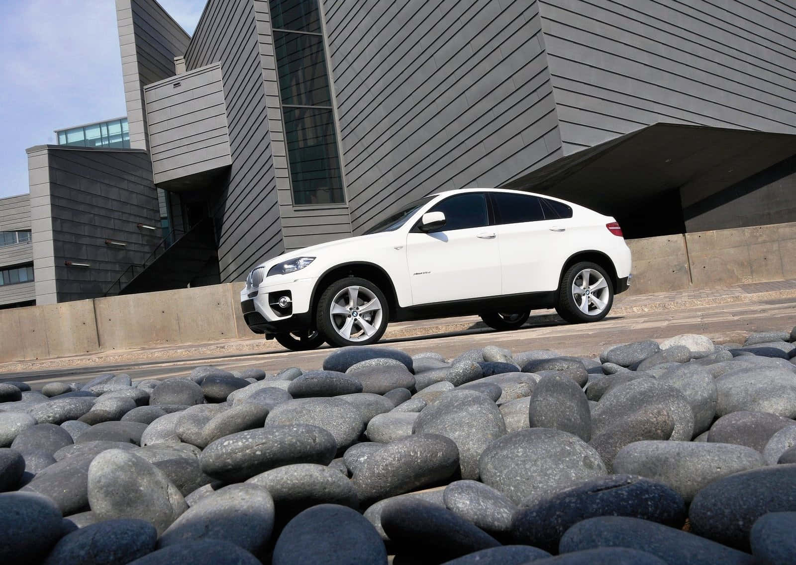 Bmw X6 桌布 壁紙 — 一輛時尚的白色BMW X6 SUV停放在現代灰色建築物旁邊的道路上，前景是光滑的灰色河石。