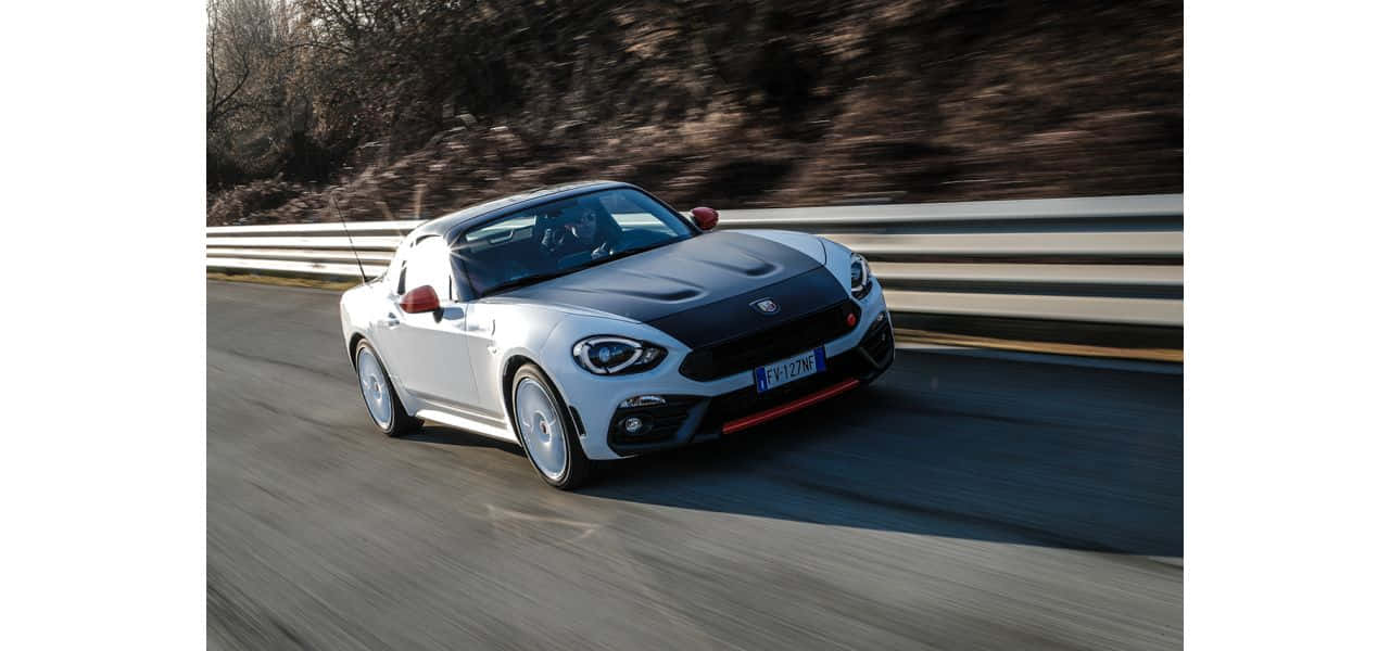 Fiat 124 Spider 桌布 壁紙 — 一台時尚的白色Fiat 124 Spider在蜿蜒的道路上疾馳，帶有動態模糊和岩石山坡背景。