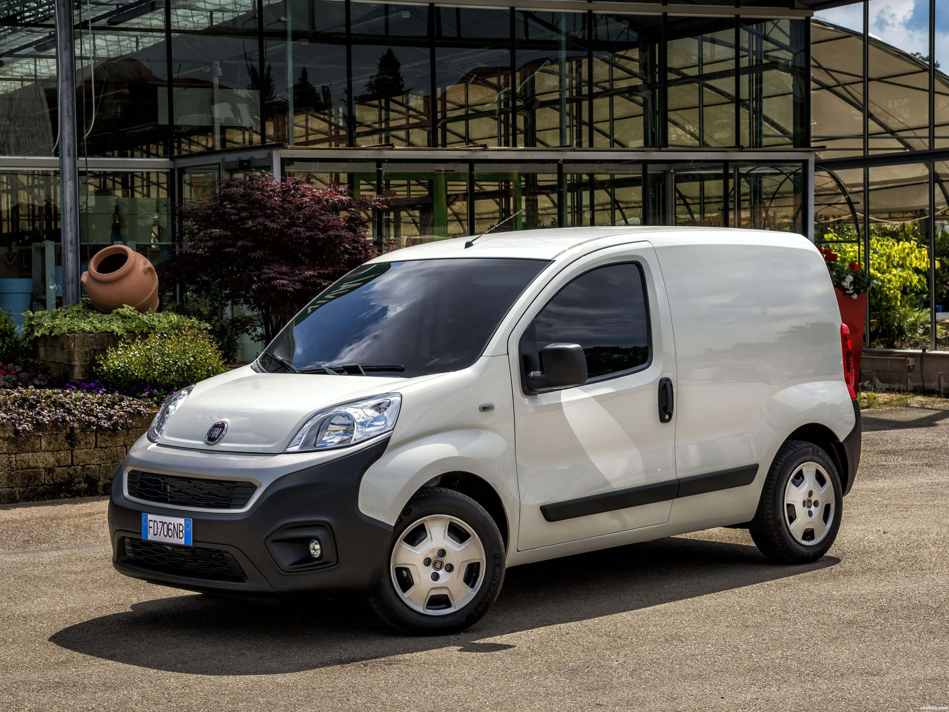 Fiat Fiorino 桌布 壁紙 — 一輛時尚的白色 Fiat Fiorino 廂型車停泊在碎石上，前面是現代玻璃溫室，周圍綠意盎然。