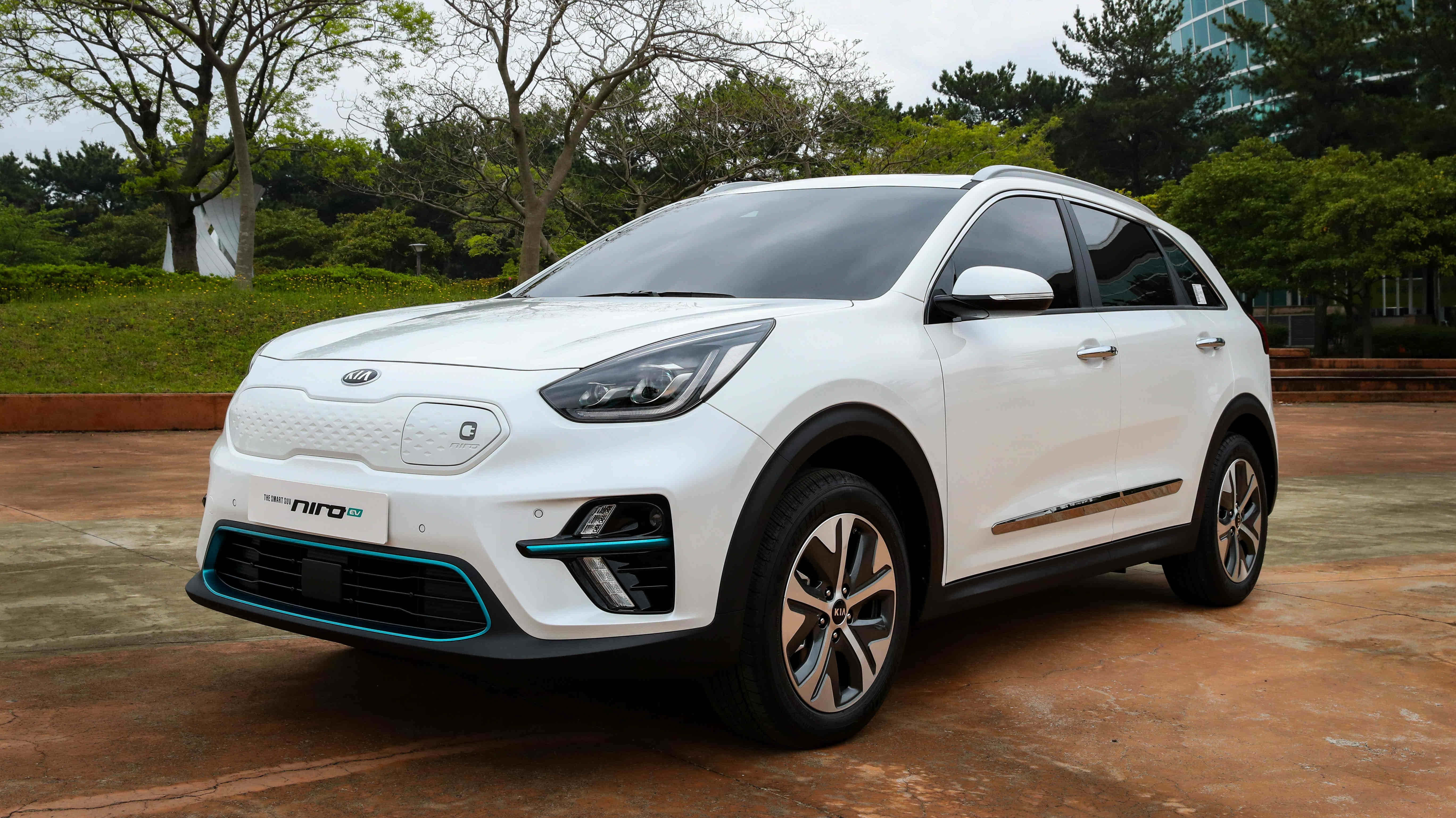 Kia Niro 桌布 壁紙 — 一輛時尚的白色Kia Niro EV SUV，帶有青綠色點綴，停在鋪砌區域，周圍環繞著綠樹和現代建築。