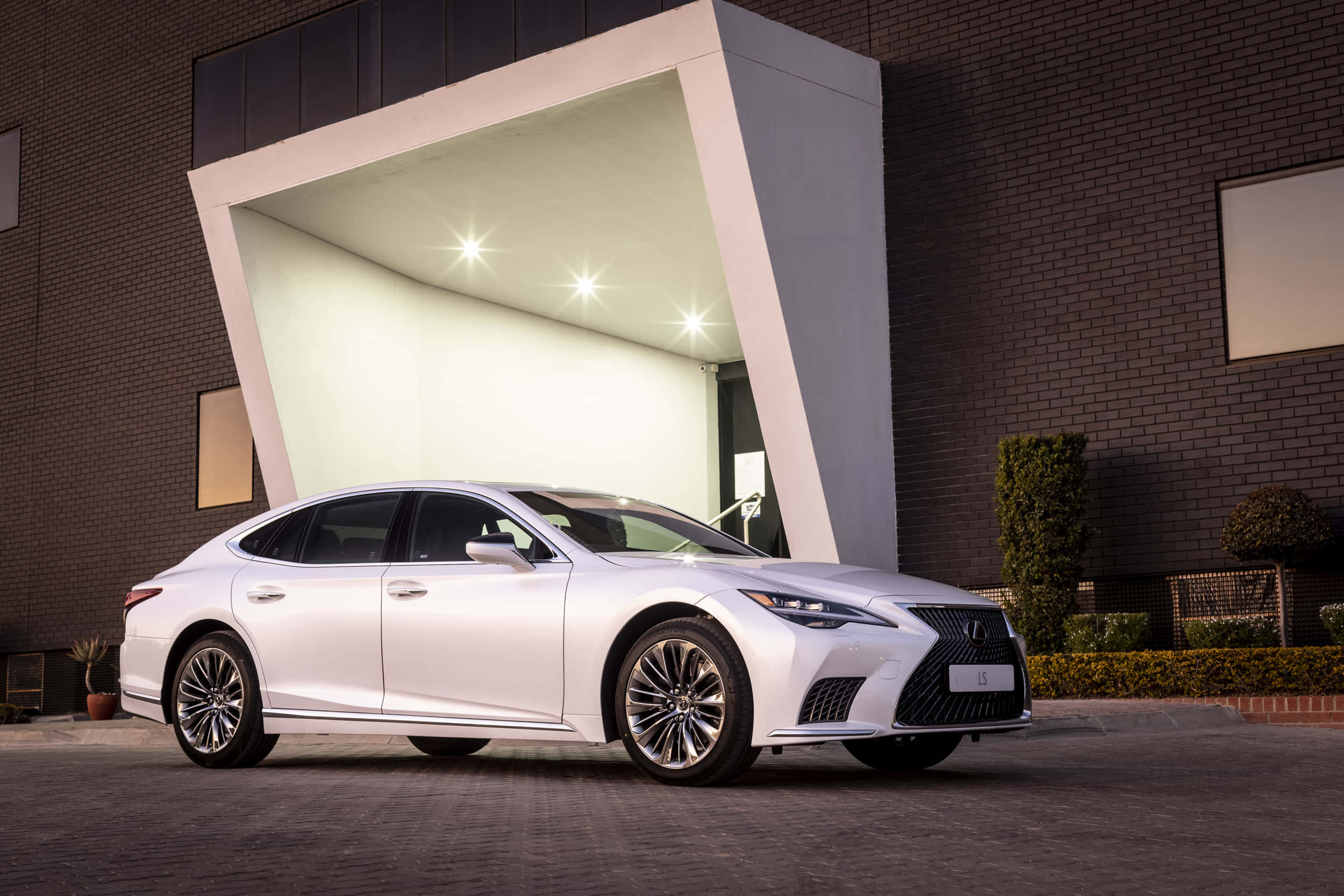 Lexus Ls 500 桌布 壁紙 — 一輛時尚的白色豪華轎車停放在現代建築簷篷下，明亮的聚光燈照亮其拋光表面。