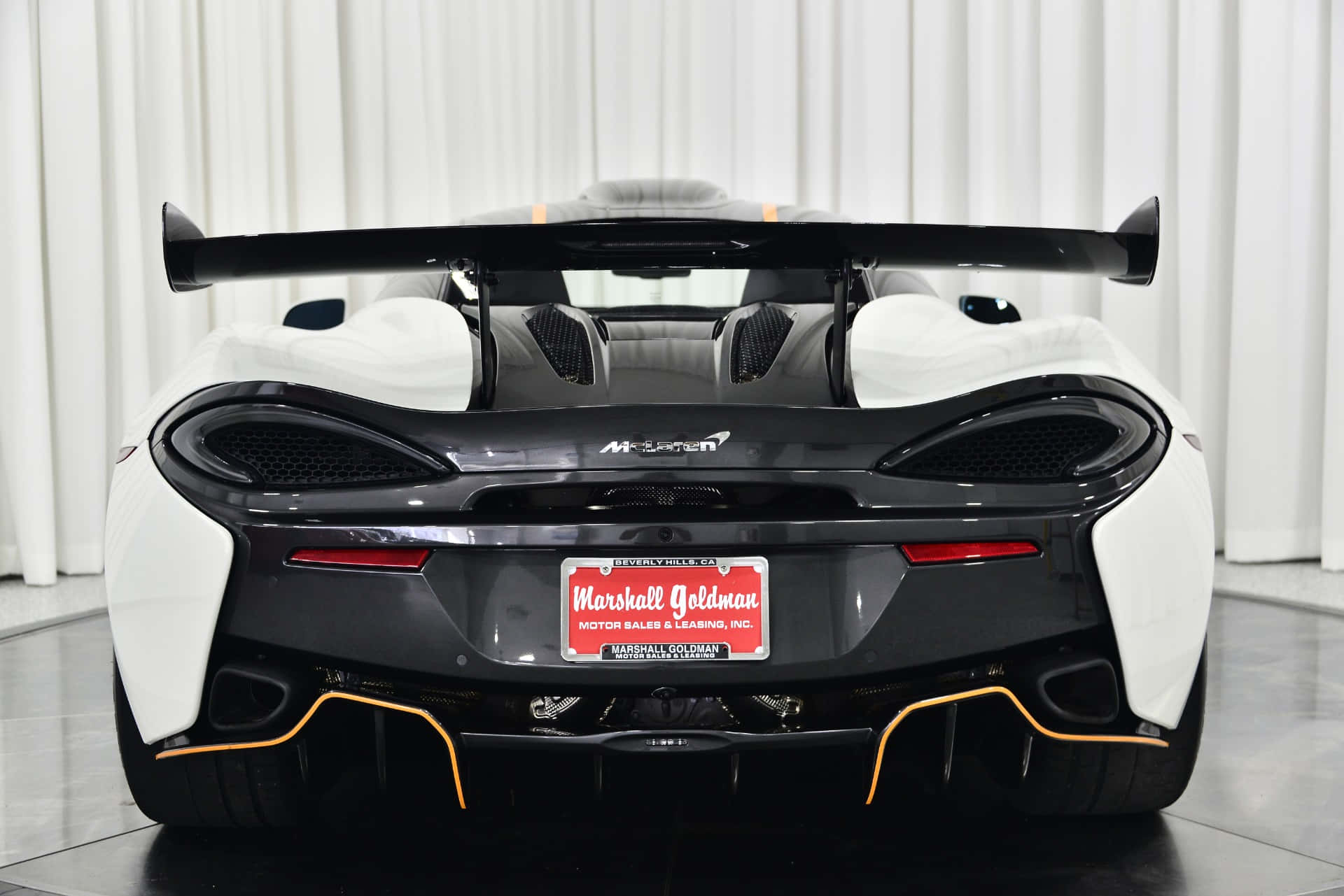 Mclaren 620r 桌布 壁紙 — 一輛時尚的白色McLaren跑車，帶有黑色飾邊和橙色飾條，在明亮的攝影棚中從後方觀看。