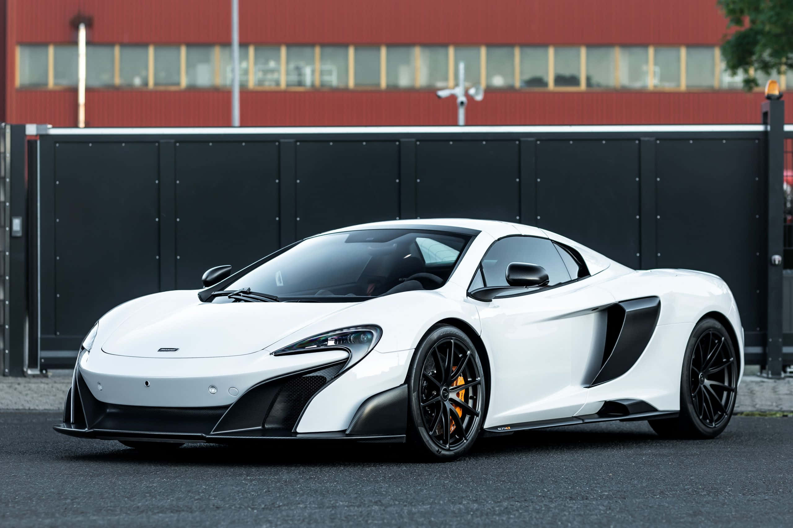 Mclaren 675lt Spider 桌布 壁紙 — 一輛時尚的白色McLaren跑車，帶有黑色飾邊和橙色制動卡鉗，停在紅樓前的瀝青路面上。
