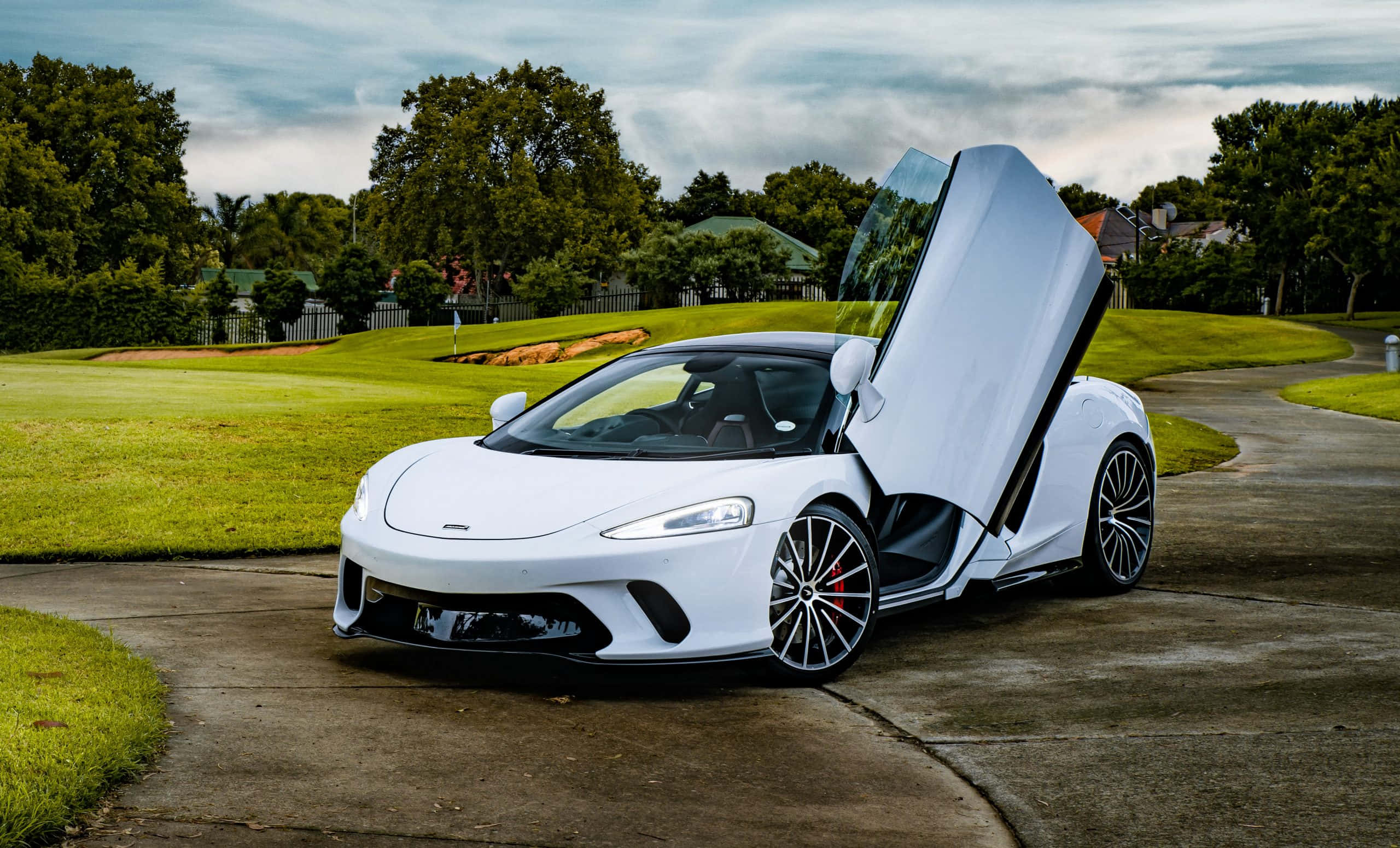 Mclaren Gt 桌布 壁紙 — 一架時尚的白色McLaren跑車，鷗翼門打開，停在鋪砌的小路上，旁邊是茂密的綠色高爾夫球場，天空多雲。
