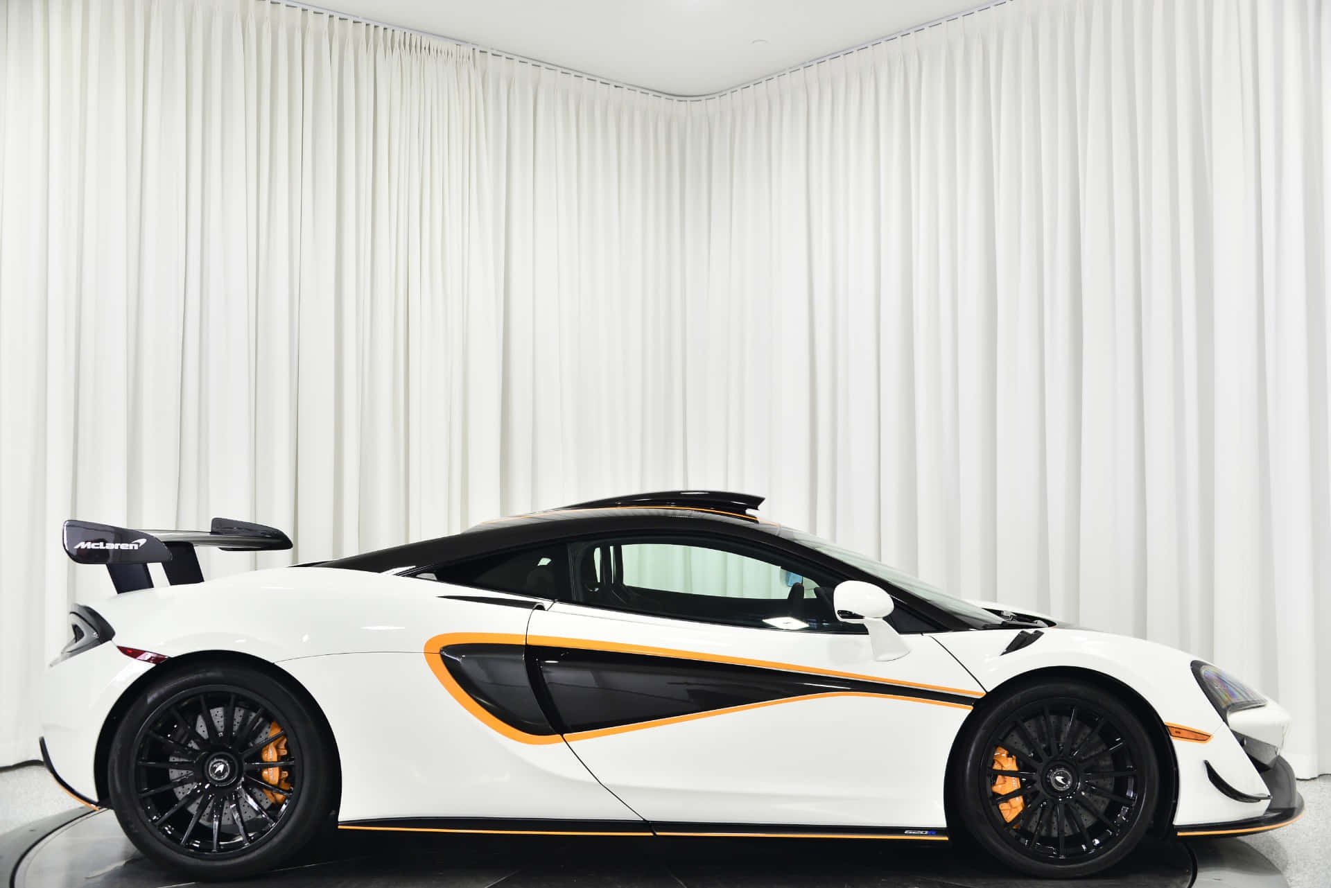 Mclaren 620r 桌布 壁紙 — 一輛時尚的白色McLaren跑車帶有橙色點綴，在純白窗簾的背景襯托下展示。