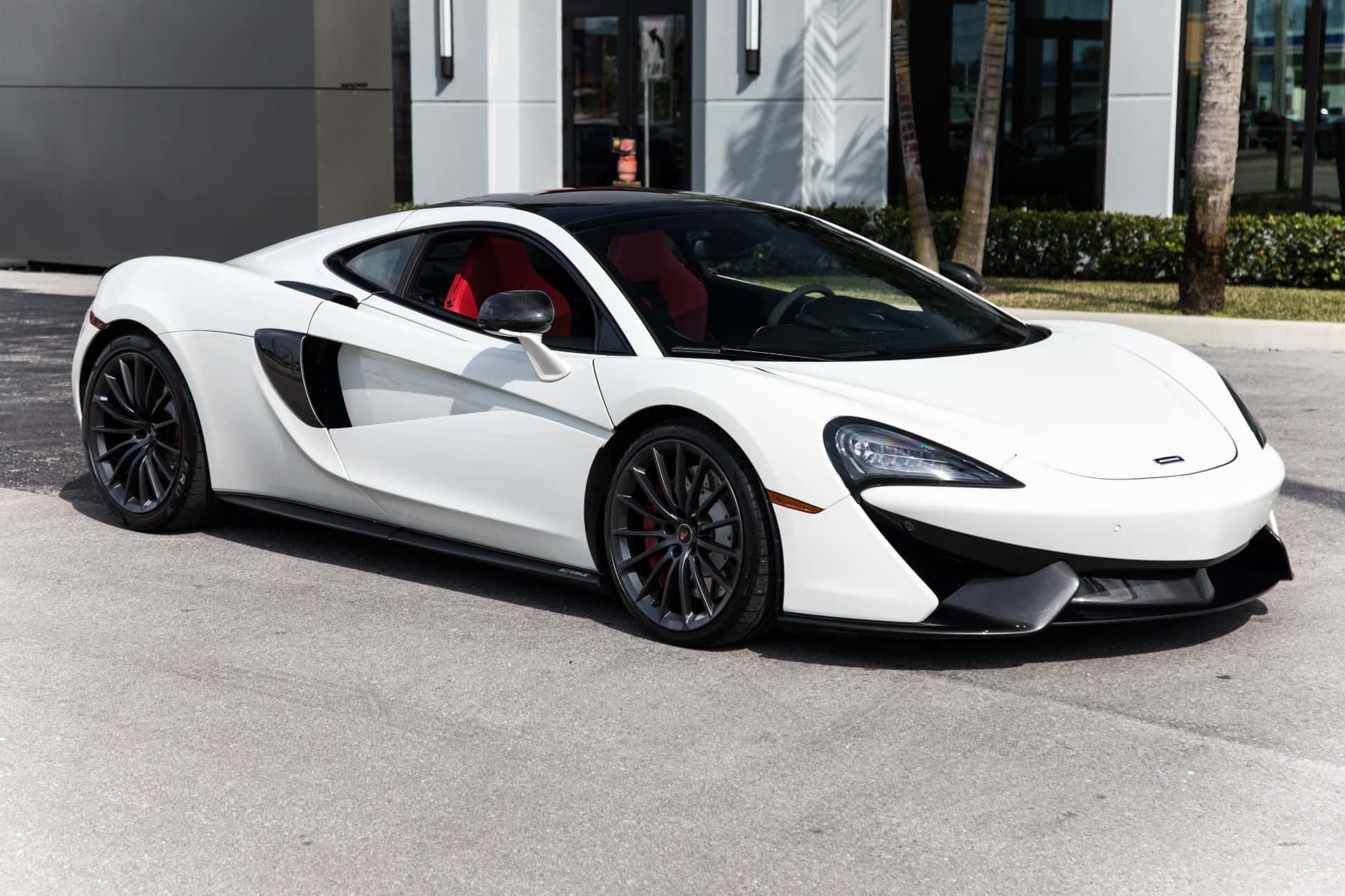 Mclaren Gt 桌布 壁紙 — 一架時尚的白色McLaren跑車，紅色內裝，停在有棕櫚樹的現代建築前的柏油路上。