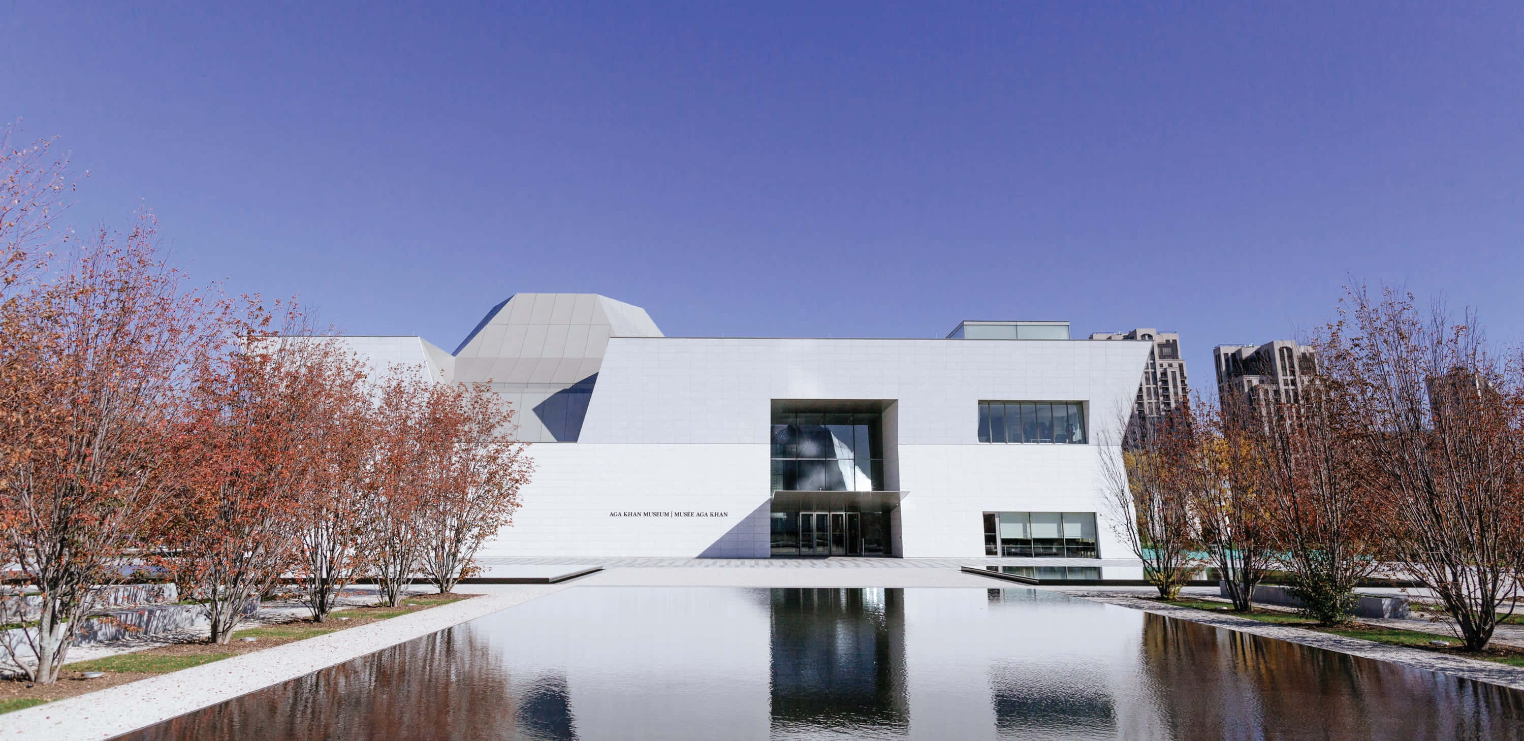 Aga Khan Museum 桌布 壁紙 — 一座時尚的白色現代博物館建築在晴朗的藍天下，映照在平靜的水景中，周圍有秋樹。