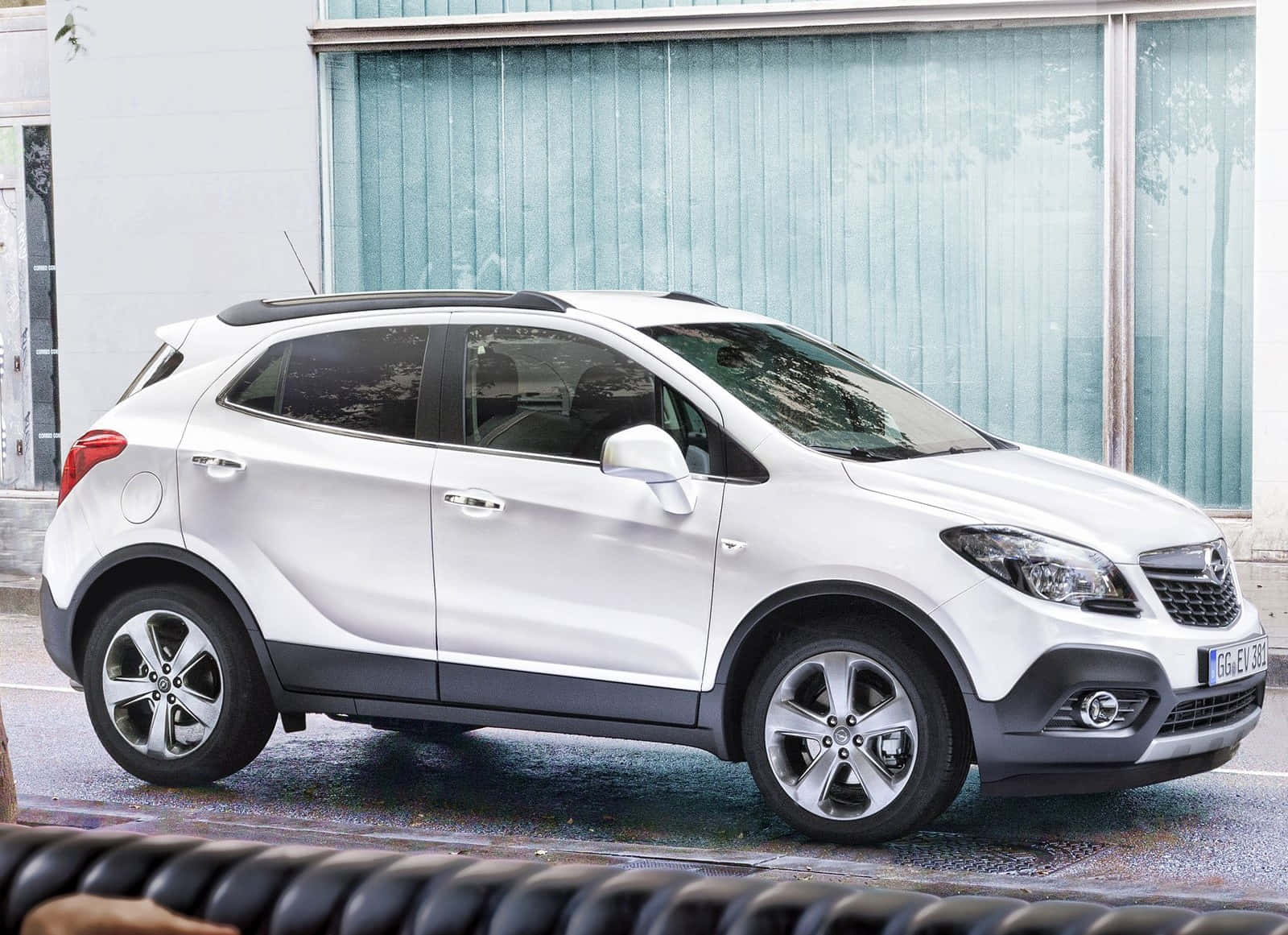 Opel Mokka 桌布 壁紙 — 一輛時尚的白色Opel Mokka SUV停放在潮濕的城市街道上，旁邊是一座帶有垂直百葉窗的現代建築。