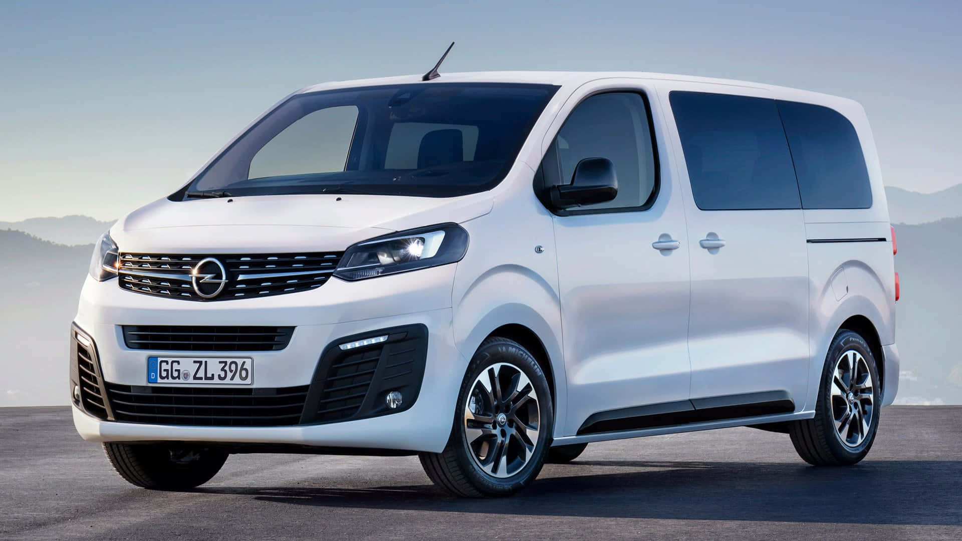 Opel Zafira 桌布 壁紙 — 一輛時尚的白色Opel Vivaro客貨車停放在鋪砌路面上，背景是朦朧的山巒，天空晴朗。