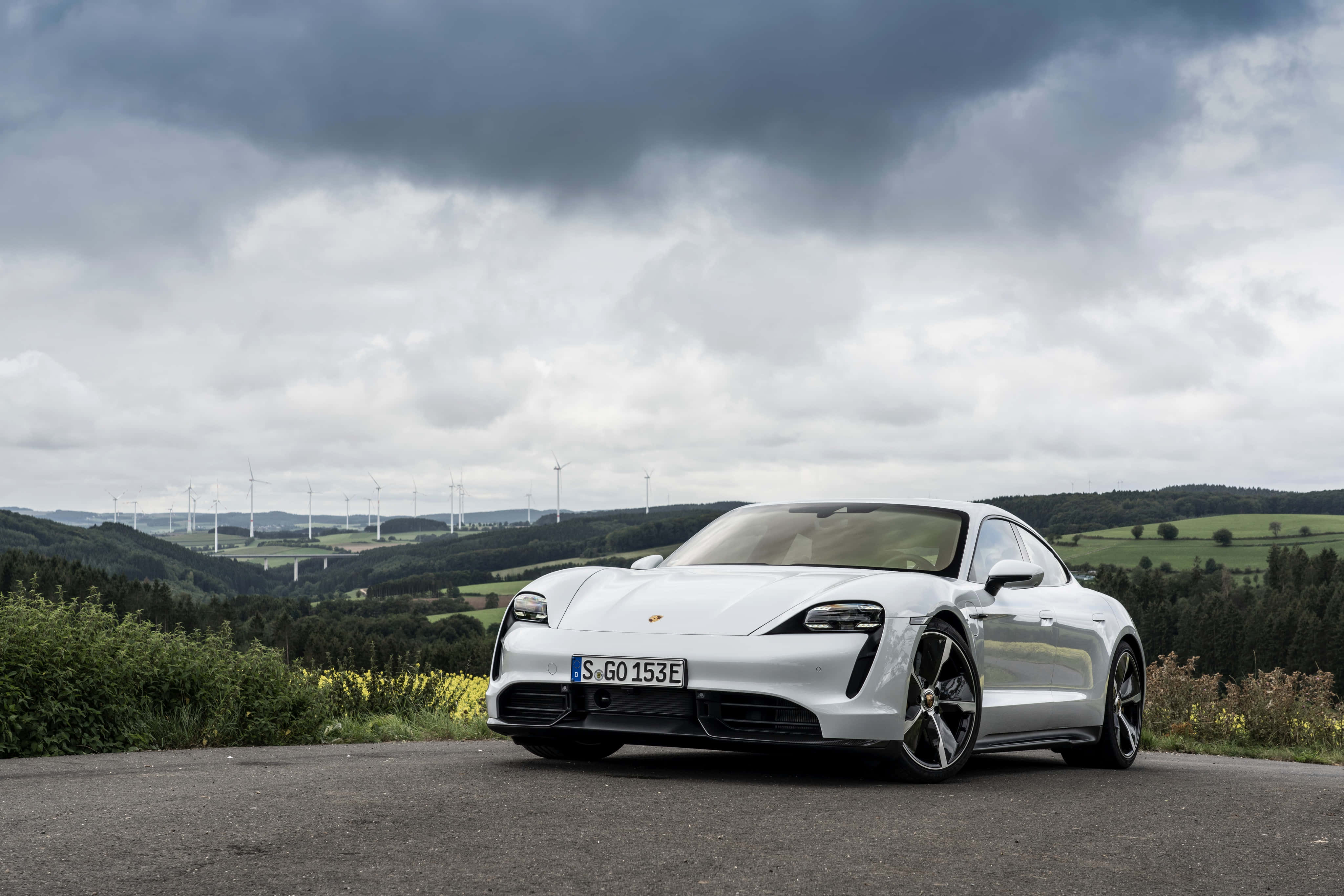 Porsche Taycan 桌布 壁紙 — 一輛時尚的白色保時捷Taycan電動車停泊在道路上，俯瞰著風力發電機下連綿起伏的綠色山丘，天空多雲。