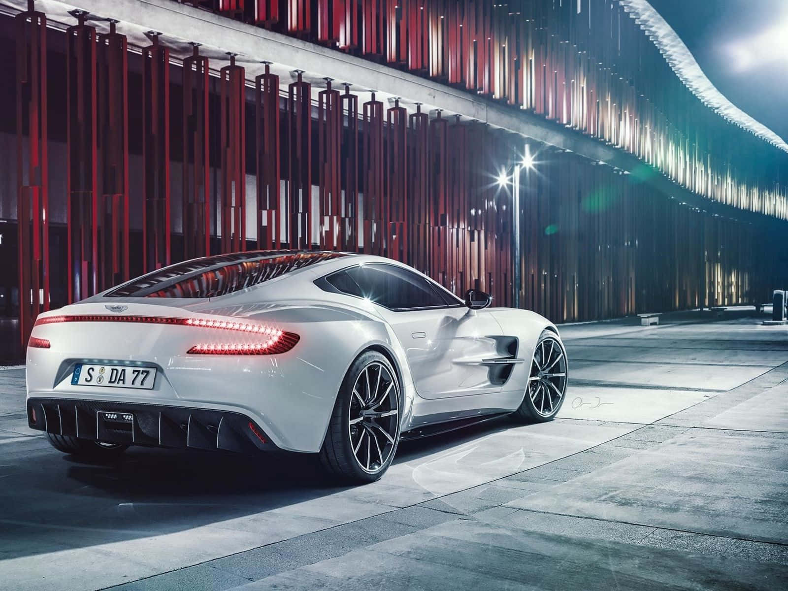 Aston Martin One-77 桌布 壁紙 — 一輛時尚的白色跑車，帶有發光的紅色尾燈，停在戲劇性的城市燈光下，背景是現代紅灰色建築。