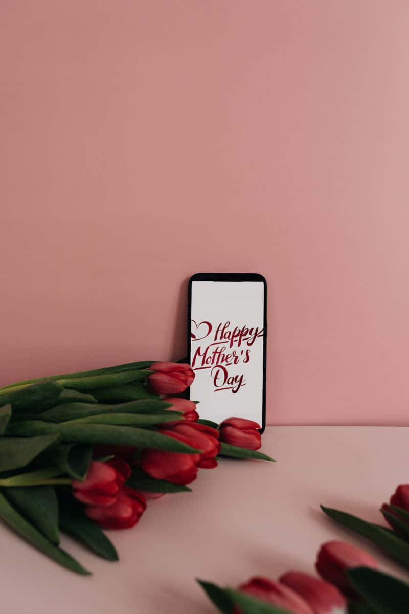  壁紙 — 一部智慧型手機顯示紅色字體的「Happy Mother's Day」，旁邊放著一束紅色鬱金香，背景是柔和的粉紅色。