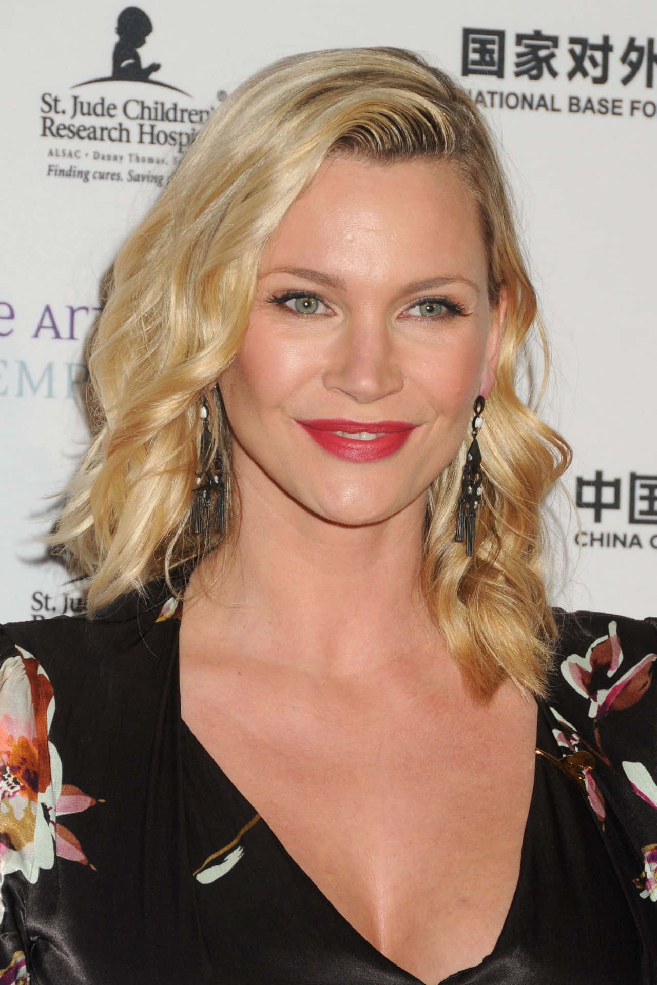 Natasha Henstridge 桌布 壁紙 — 一位微笑的金髮女性，塗著紅色口紅，穿著花卉襯衫，在品牌背景前於慈善活動中擺姿勢。