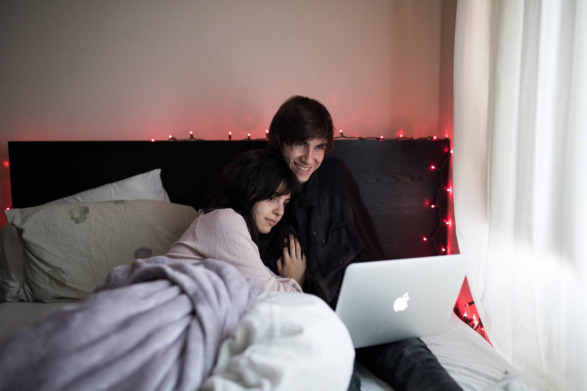 一對情侶在床上依偎，看著身後有紅色串燈發光的筆記型電腦。 — Macbook Pro 壁紙
