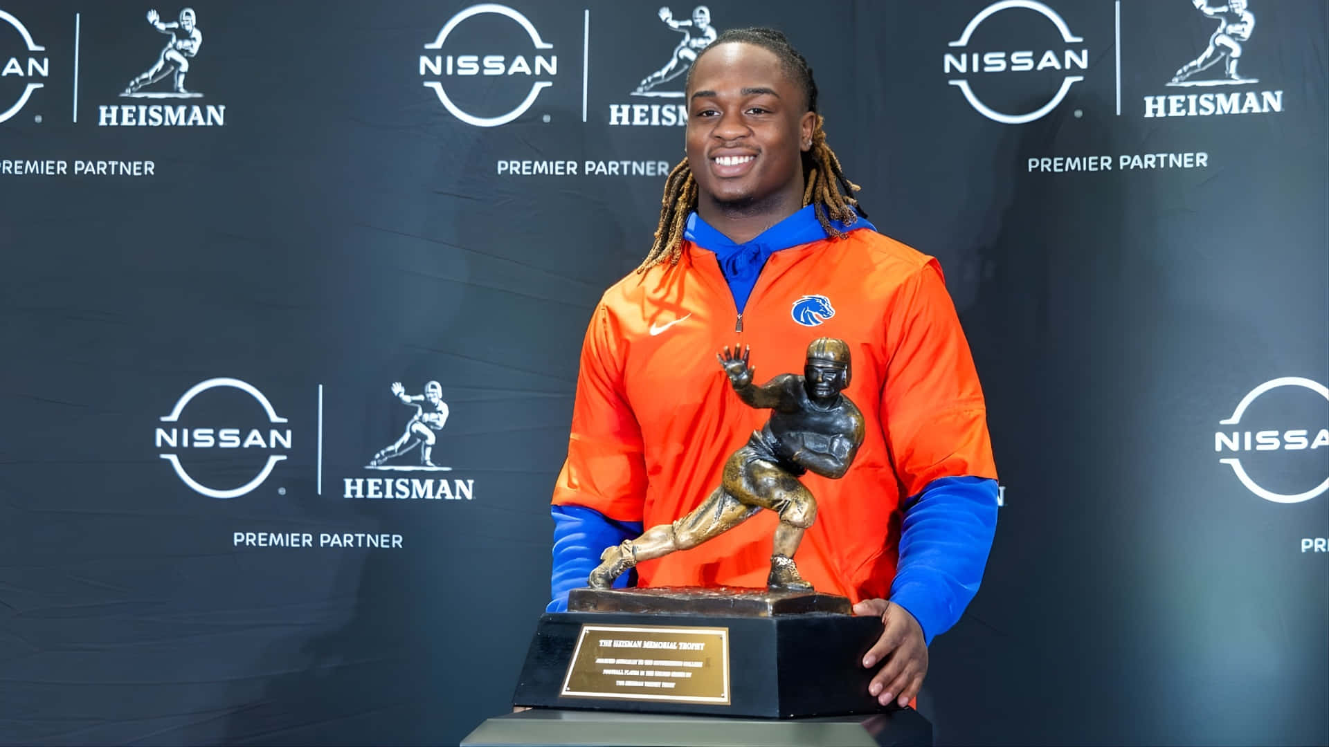  壁紙 — 一位面帶微笑的美式足球員，身穿橙色和藍色外套，在品牌背景前自豪地高舉海斯曼獎盃。