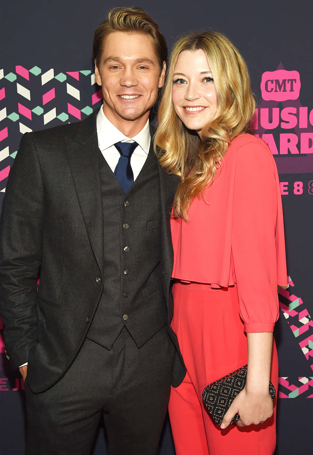 Sarah Roemer 桌布 壁紙 — 一位微笑的男士穿著深色西裝，一位女士穿著珊瑚色連衣裙，在色彩繽紛的CMT音樂獎背景前合影。