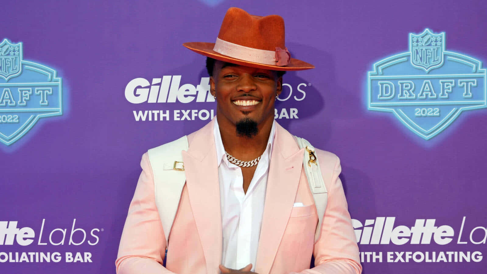  壁紙 — 一位微笑的男子身穿粉紅色西裝和橙色帽子，在2022年NFL選秀活動中，以紫色品牌背景自信地擺姿勢。