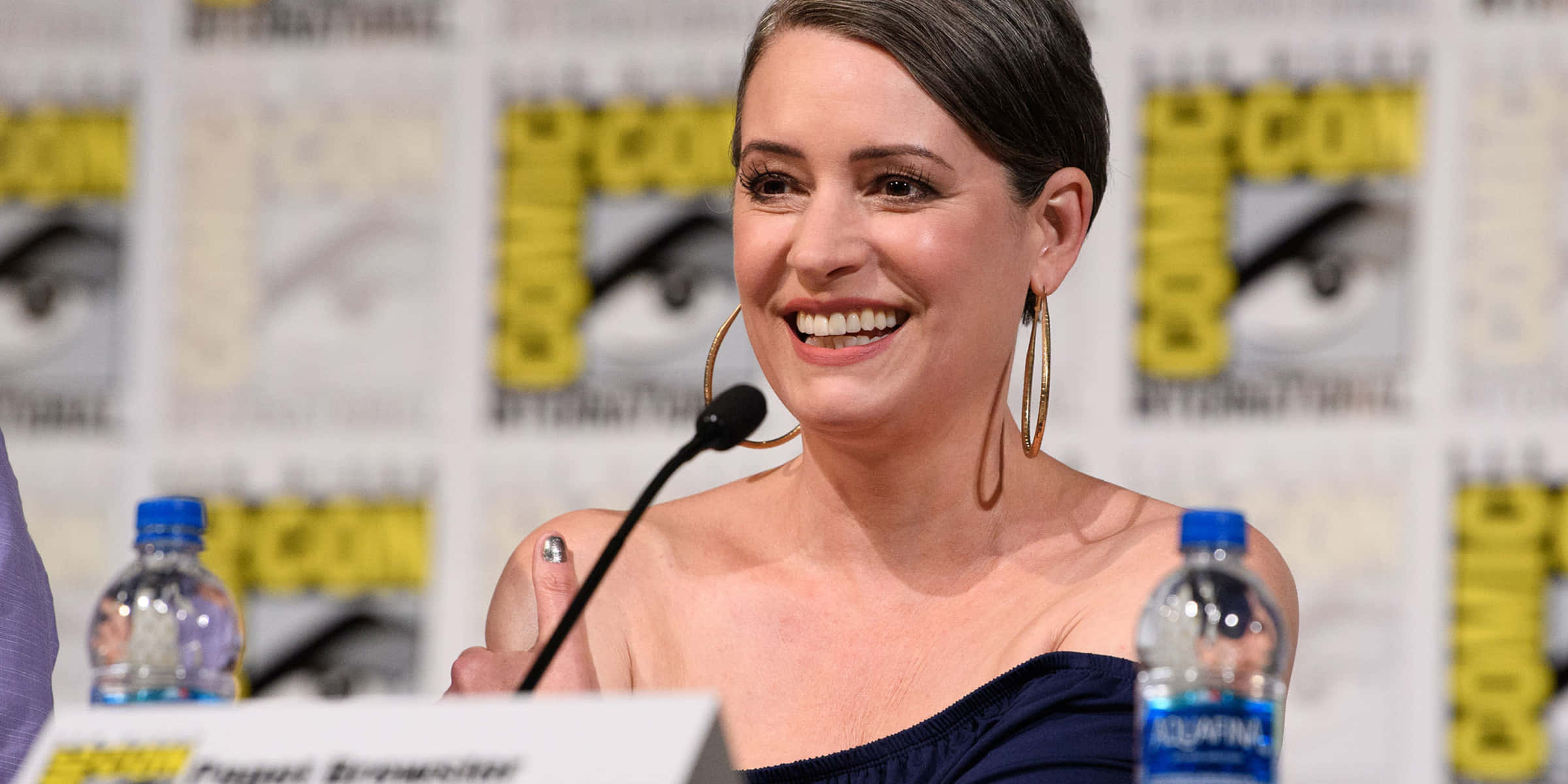 paget brewster 桌布 壁紙 — 一位微笑的短黑髮女性戴著金色耳環，在帶有品牌背景和水瓶的Comic-Con座談會上發言。