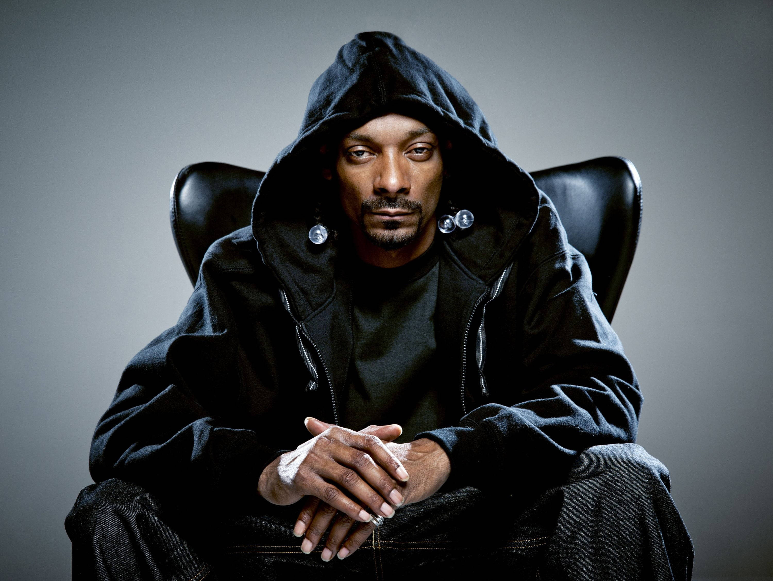 Snoop 桌布 壁紙 — Snoop Dogg 冷靜地坐在黑色皮椅上，身穿黑色連帽衫，戴著銀色耳環，手部纏著繃帶。
