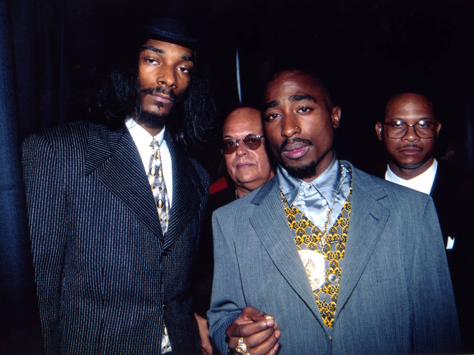 桌布 壁紙 — Snoop Dogg 和 Tupac Shakur 身穿筆挺西裝，在深色背景前自信地擺姿勢，身後還有另外三名男子。